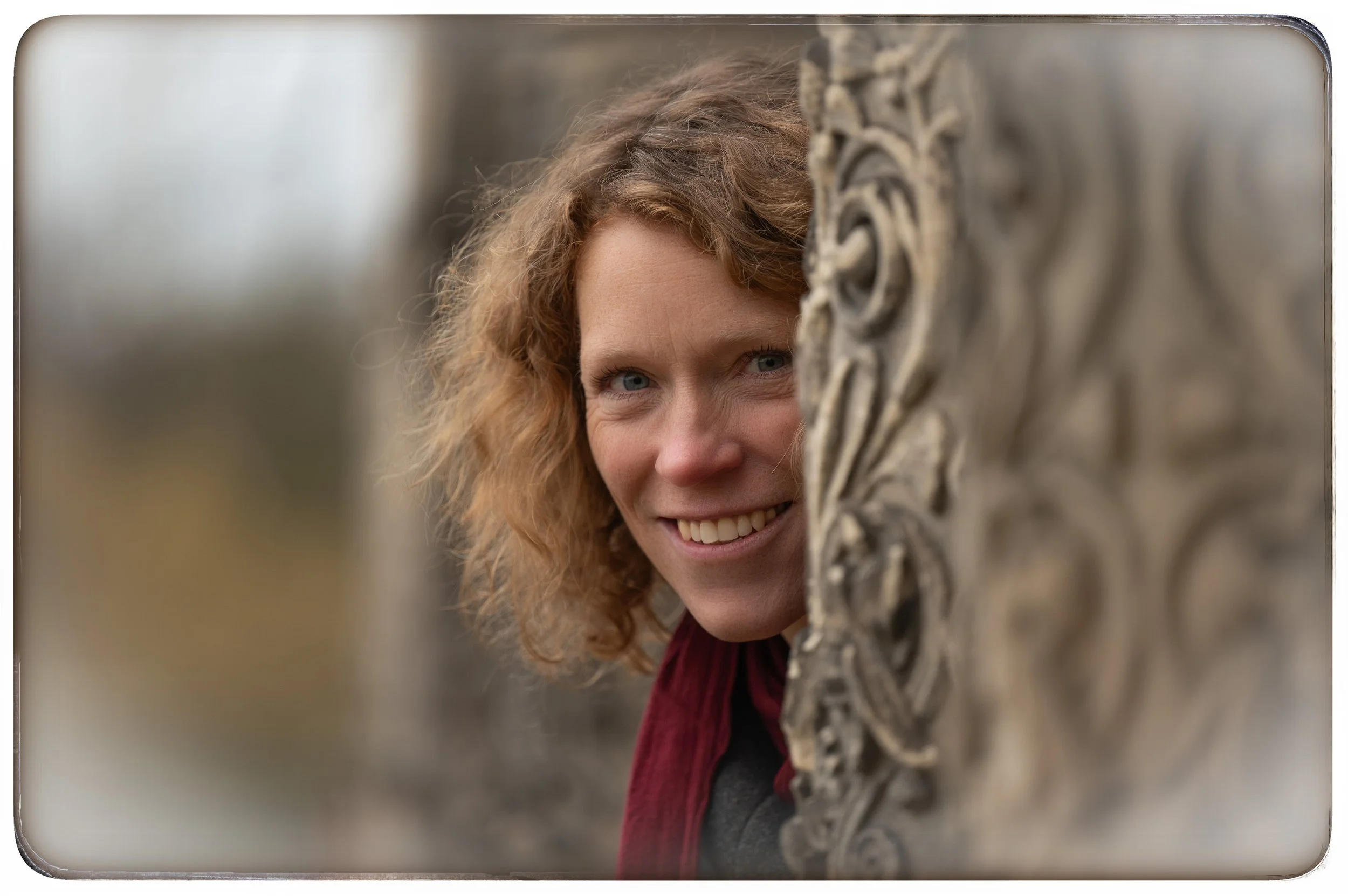 Eine Frau mit lockigem, rotem Haar, die hinter einem verzierten Stein ornament lauert und in die Kamera lächelt.