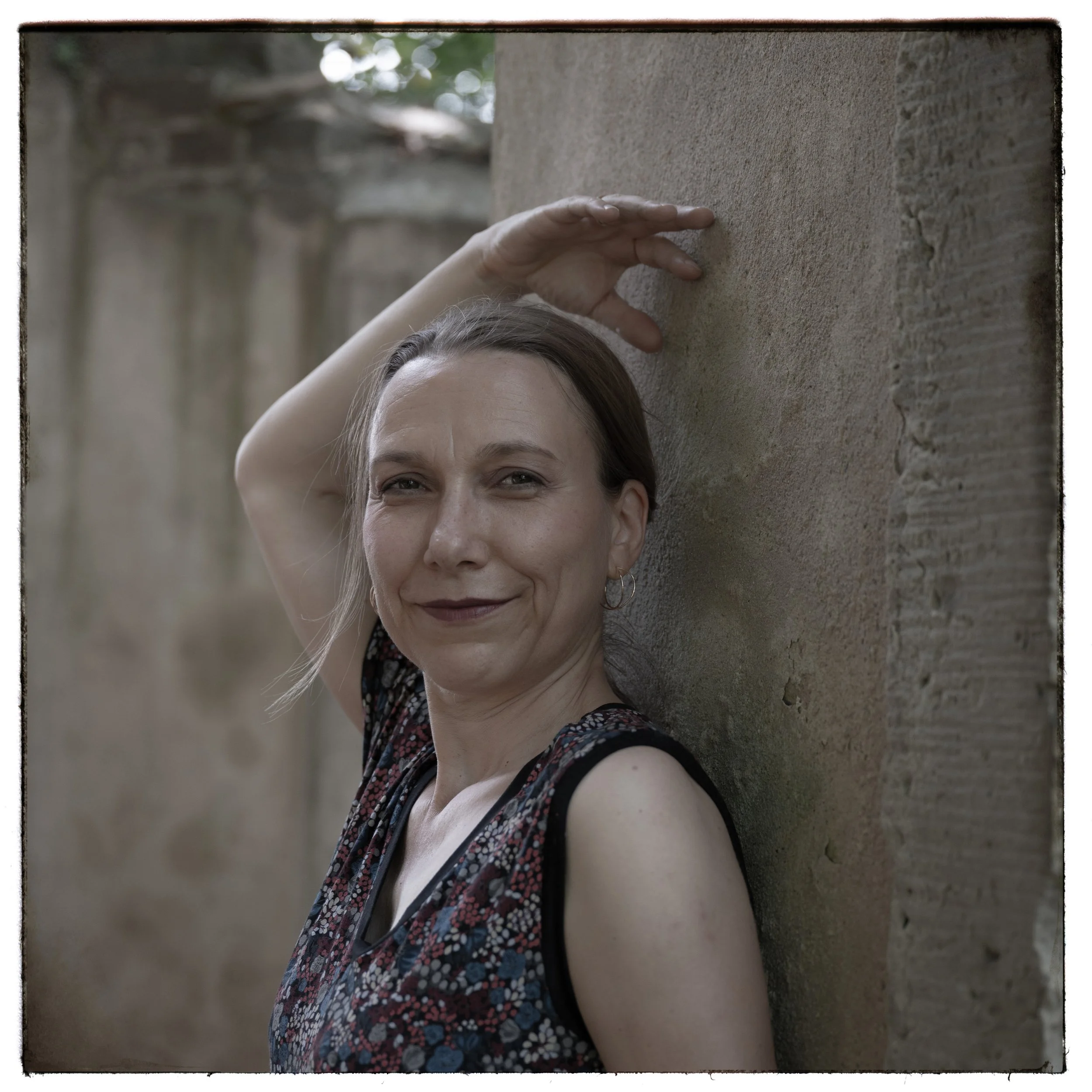 Eine Frau lehnt an einer Sandsteinmauer, lächelnd, mit dunklem Lippenstift und einem schwarzen Kleid mit buntem Muster, im Hintergrund unscharf