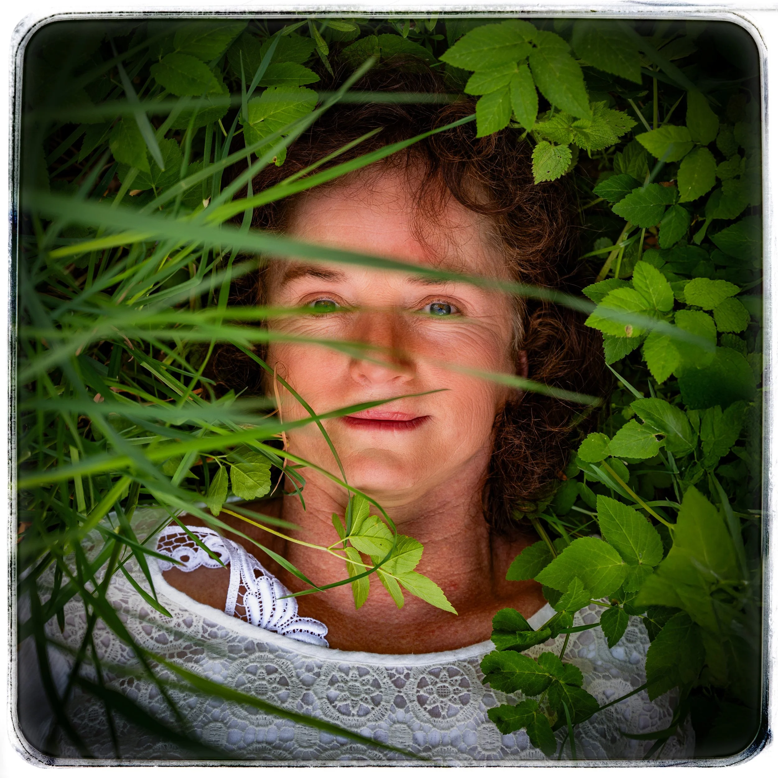 Frau liegt im Grünen, umgeben von Blättern und Pflanzen, fotografiert von oben, trägt ein weißes Spitzenoberteil, lächelt leicht, mit braunen, lockigen Haaren.