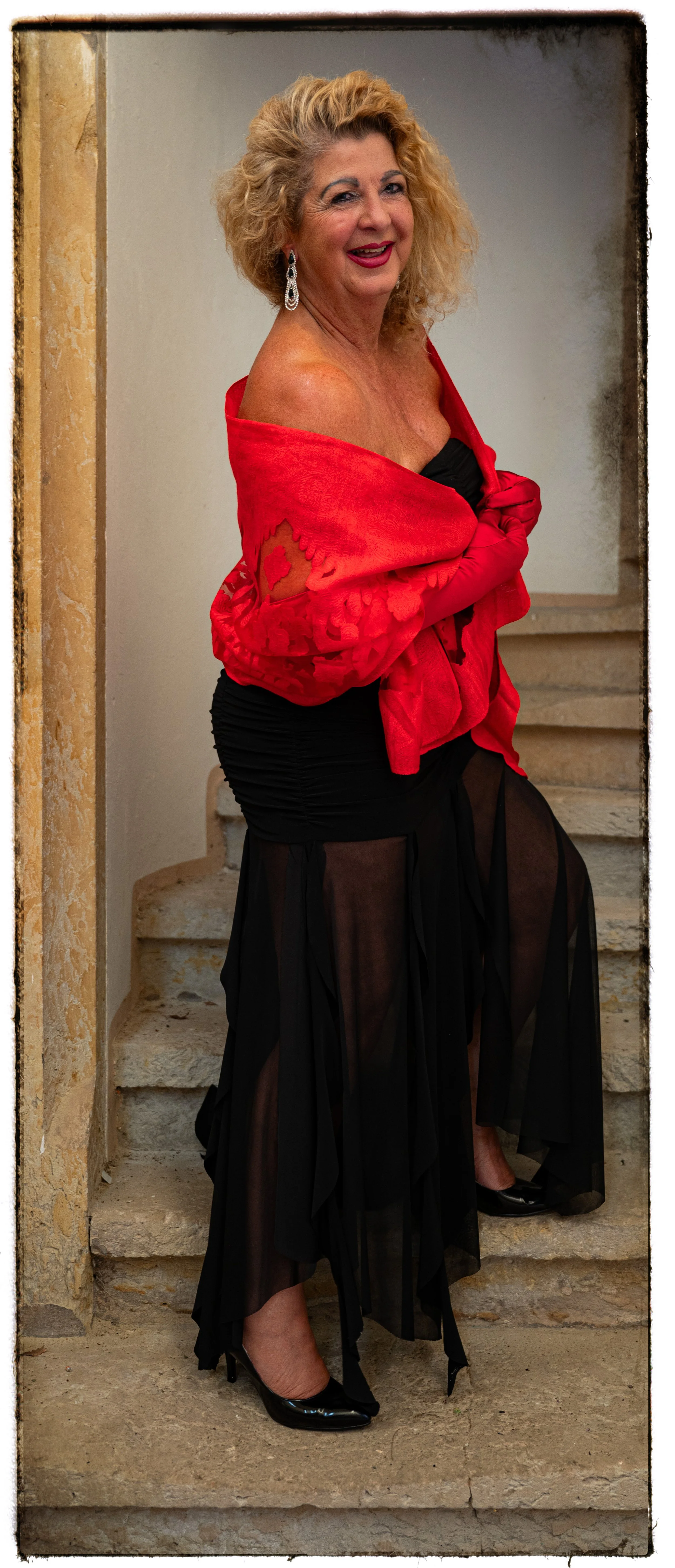 Eine älteren Frau mit lockigem blonden Haar in einem roten Abendkleid und schwarzen transparenten Rock auf Steinstufen stehend, lächelnd, bei einer Veranstaltung.