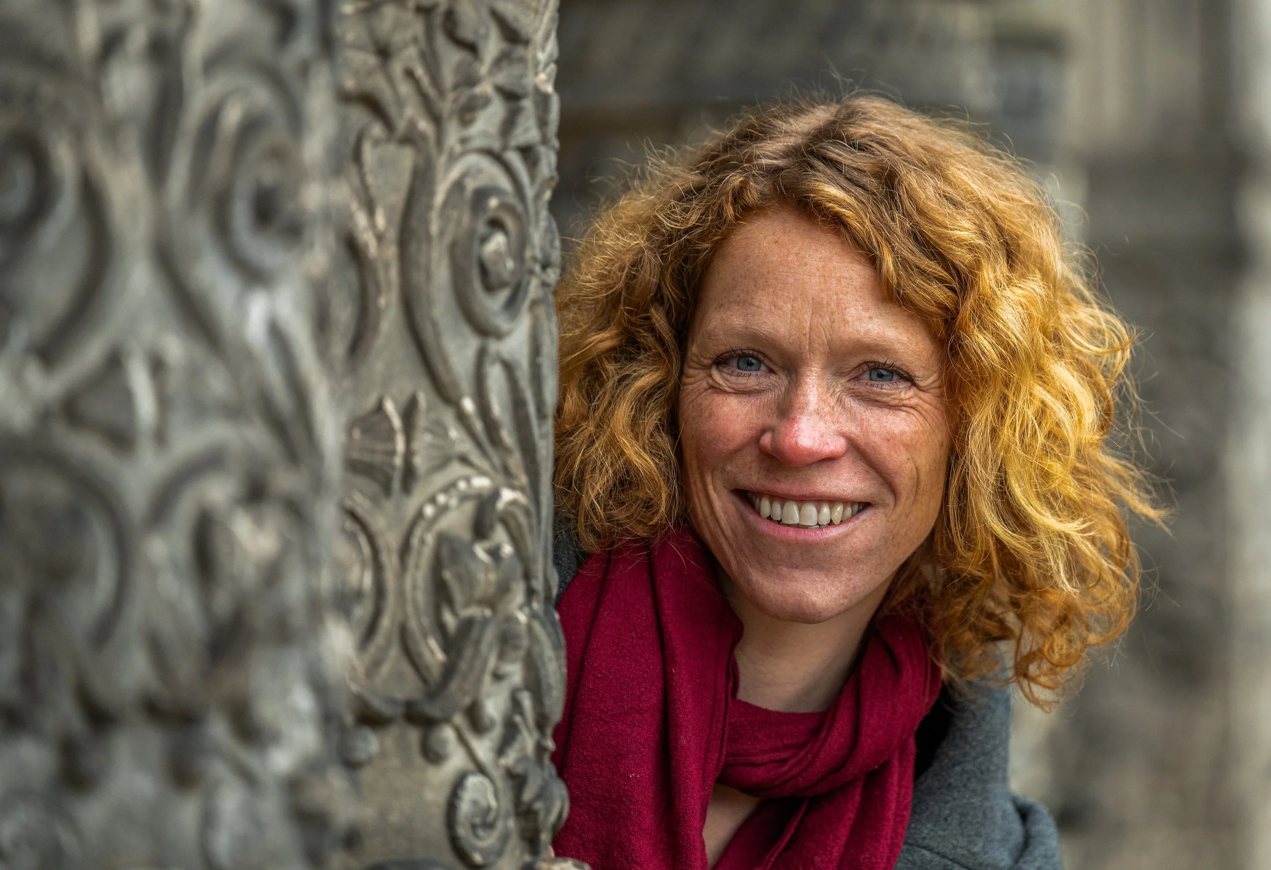 Eine Frau mit roten, lockigen Haaren lächelt beim Posieren neben einer verzierten Säule aus Stein.