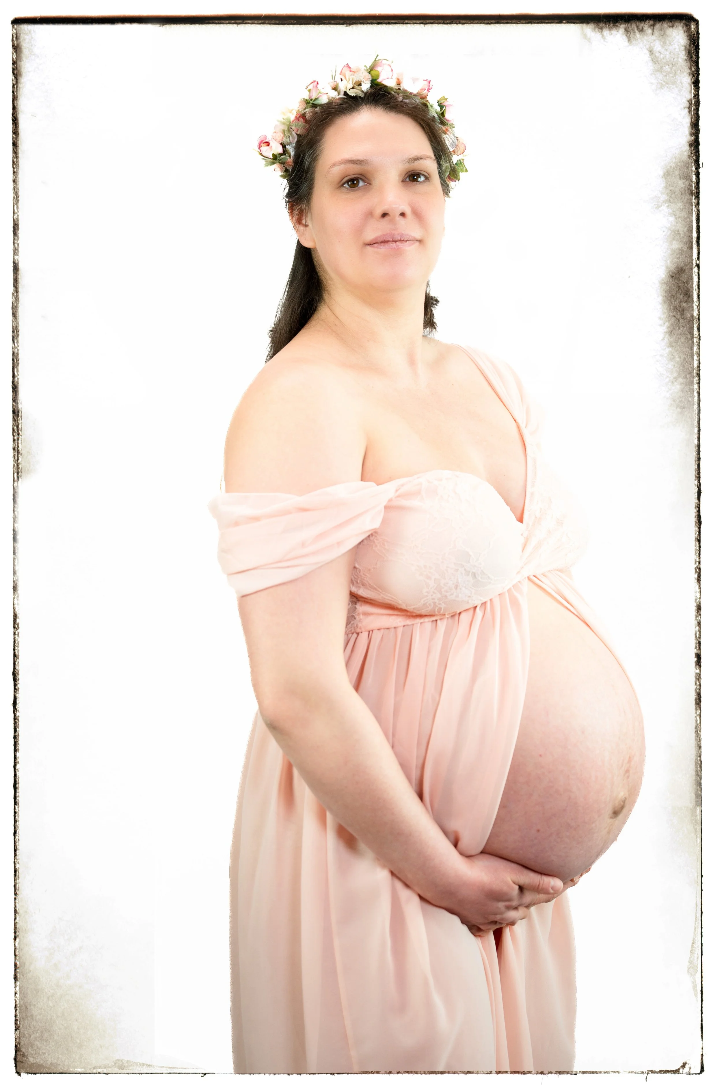 schwangere Frau trägt ein rosa Kleid und einen Blumenkranz auf dem Kopf, steht vor weißem Hintergrund
