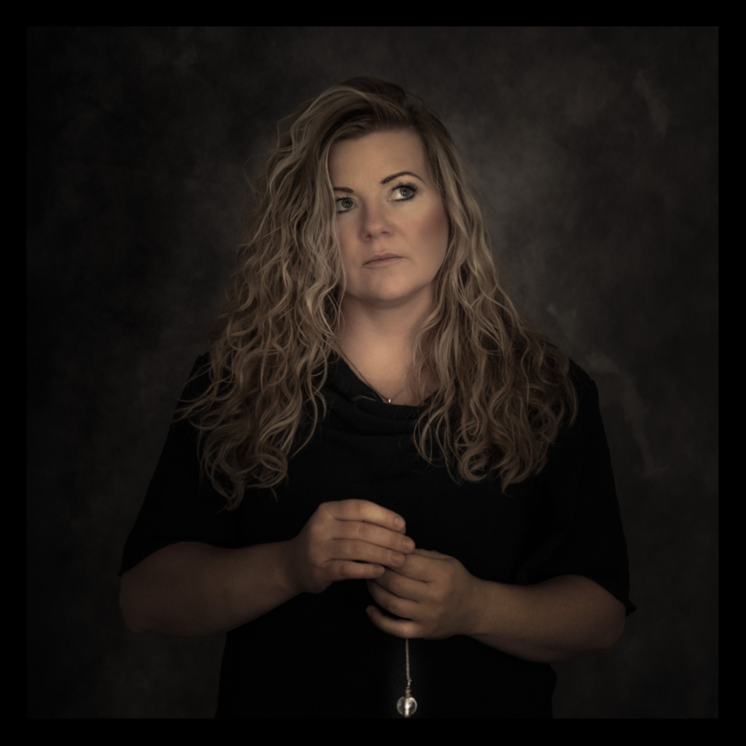 Porträt einer Frau mit langen, lockigen, hellbraunen Haaren, schwarz gekleidet, vor dunklem Hintergrund, blickt nach rechts, nachdenklich.