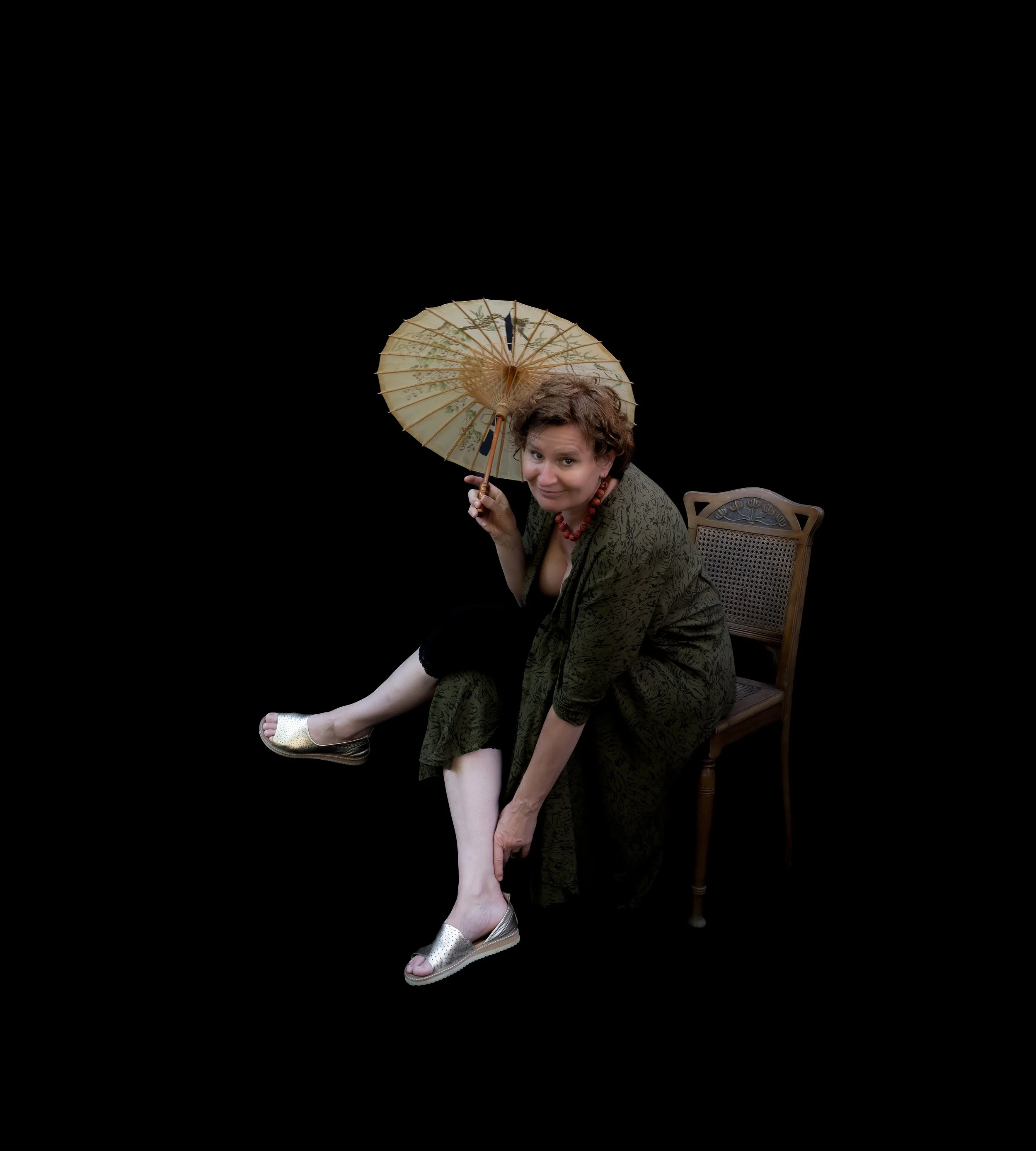 Eine Frau sitzt auf einem Stuhl, trägt einen grünen Mantel, zeigt auf ihren Fuß, hält einen laternenförmigen Regenschirm und blickt humorvoll. Hintergrund ist schwarz.