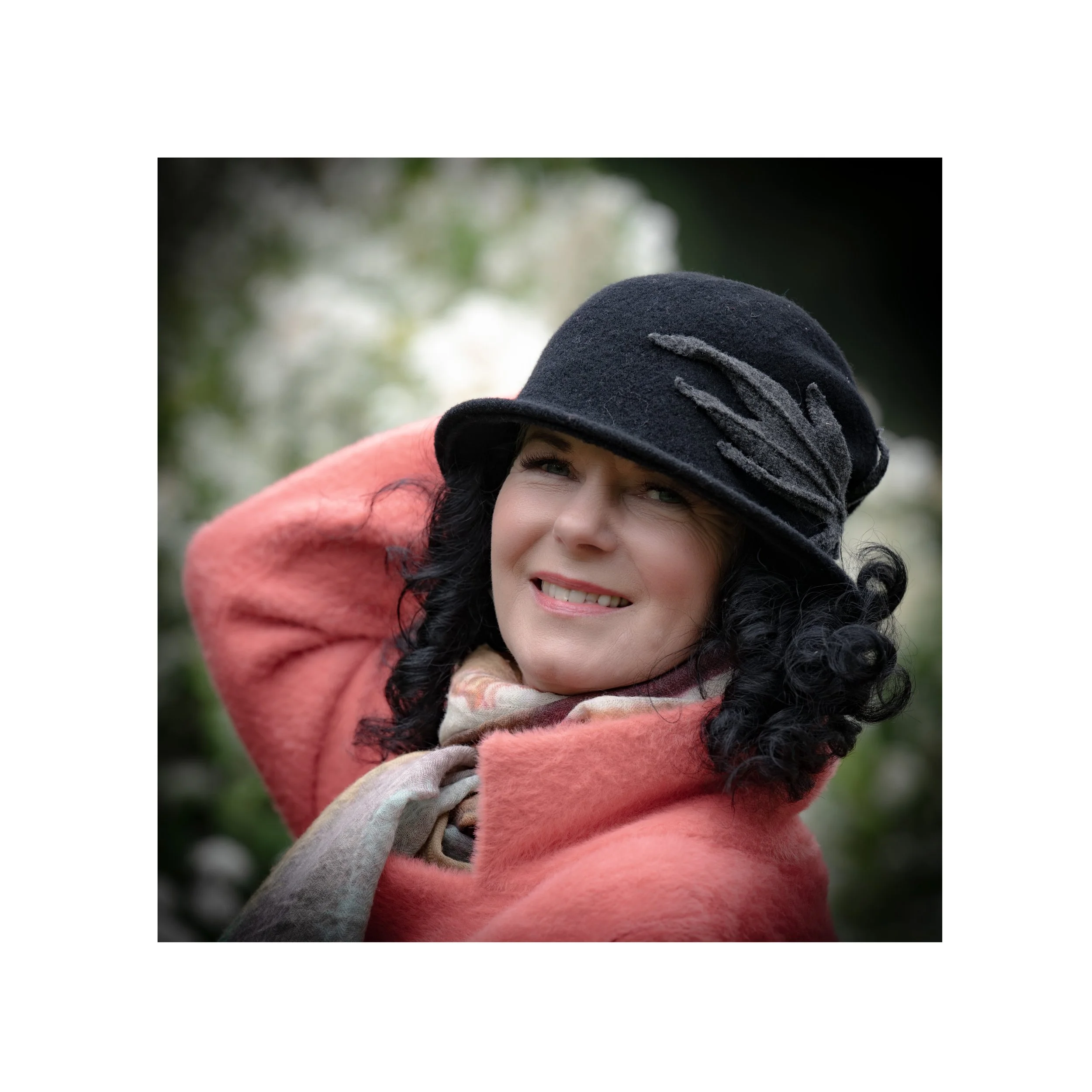 Eine Frau trägt einen schwarzen Hut mit verzerrtem Design, einen pinken Mantel und einen beigen Schal. Sie hat dunkle, lockige Haare und lächelt bei sonnigem Wetter im Freien.