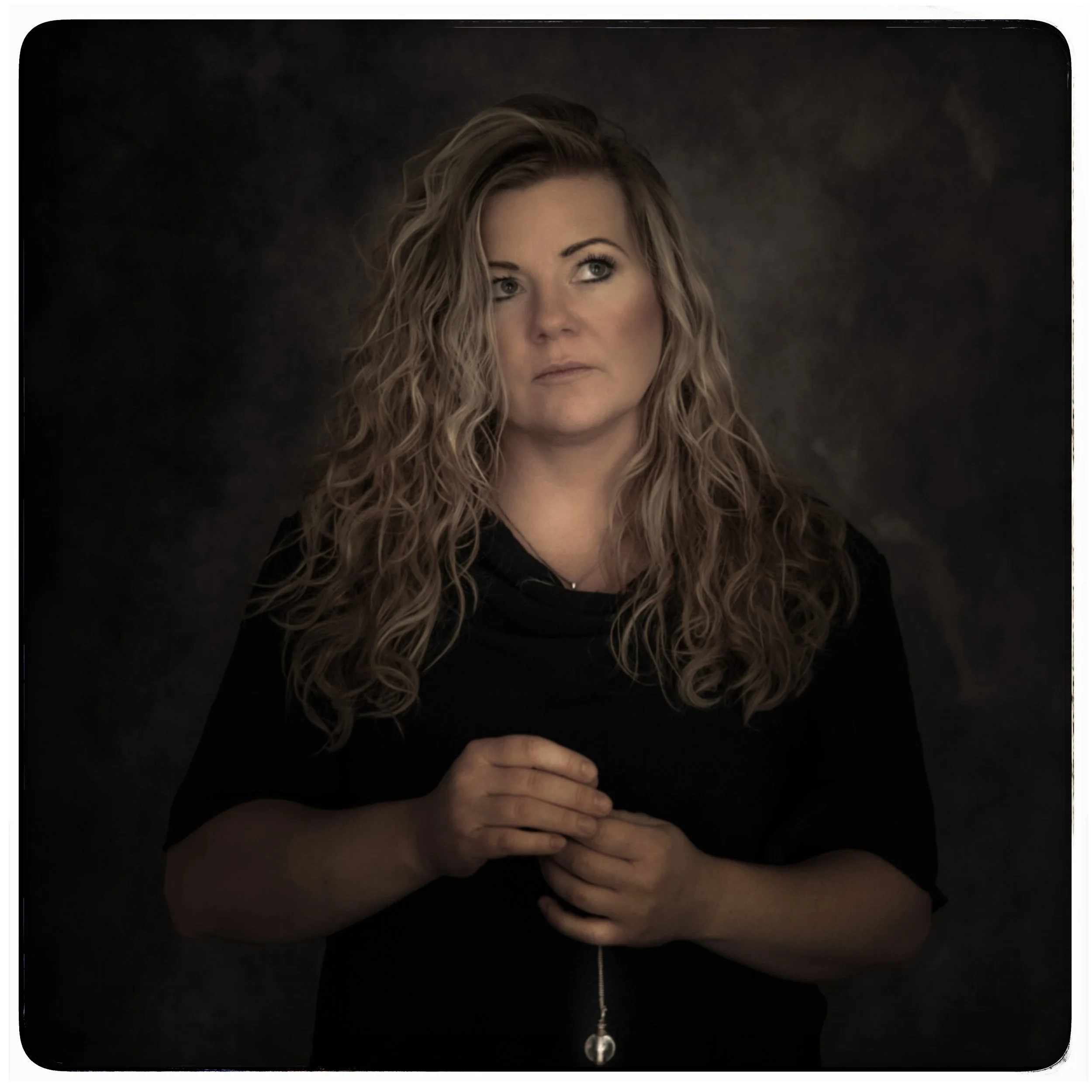 Eine Frau mit lockigem, hellbraunem Haar trägt einen schwarzen Pullover und hält eine Halskette mit einem Anhänger, Blick nach rechts, dunkler Hintergrund.