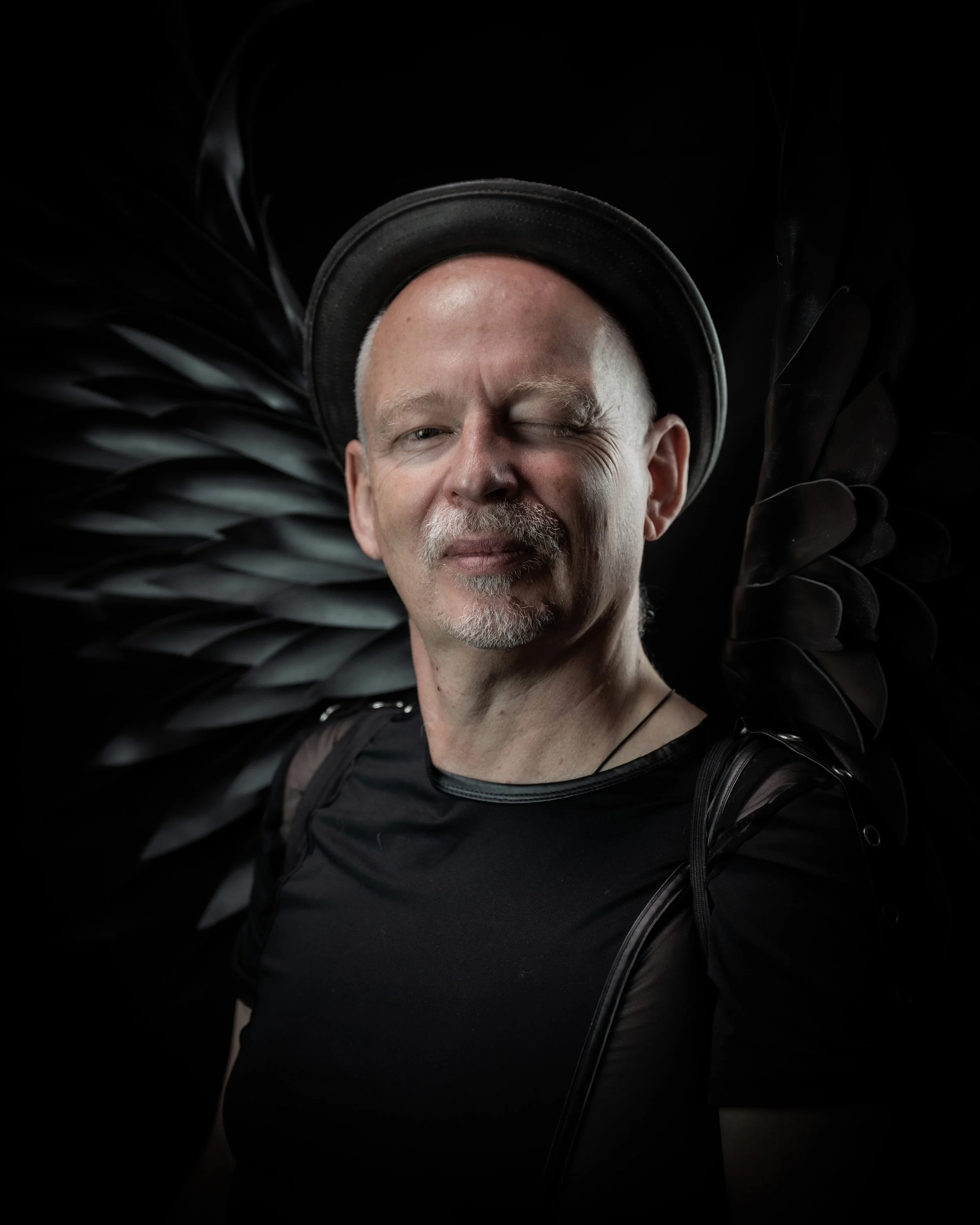 Mann mit schwarzem Hut und schwarzen Flügeln vor schwarzem Hintergrund, eines Auges geschlossen, lächelnd.