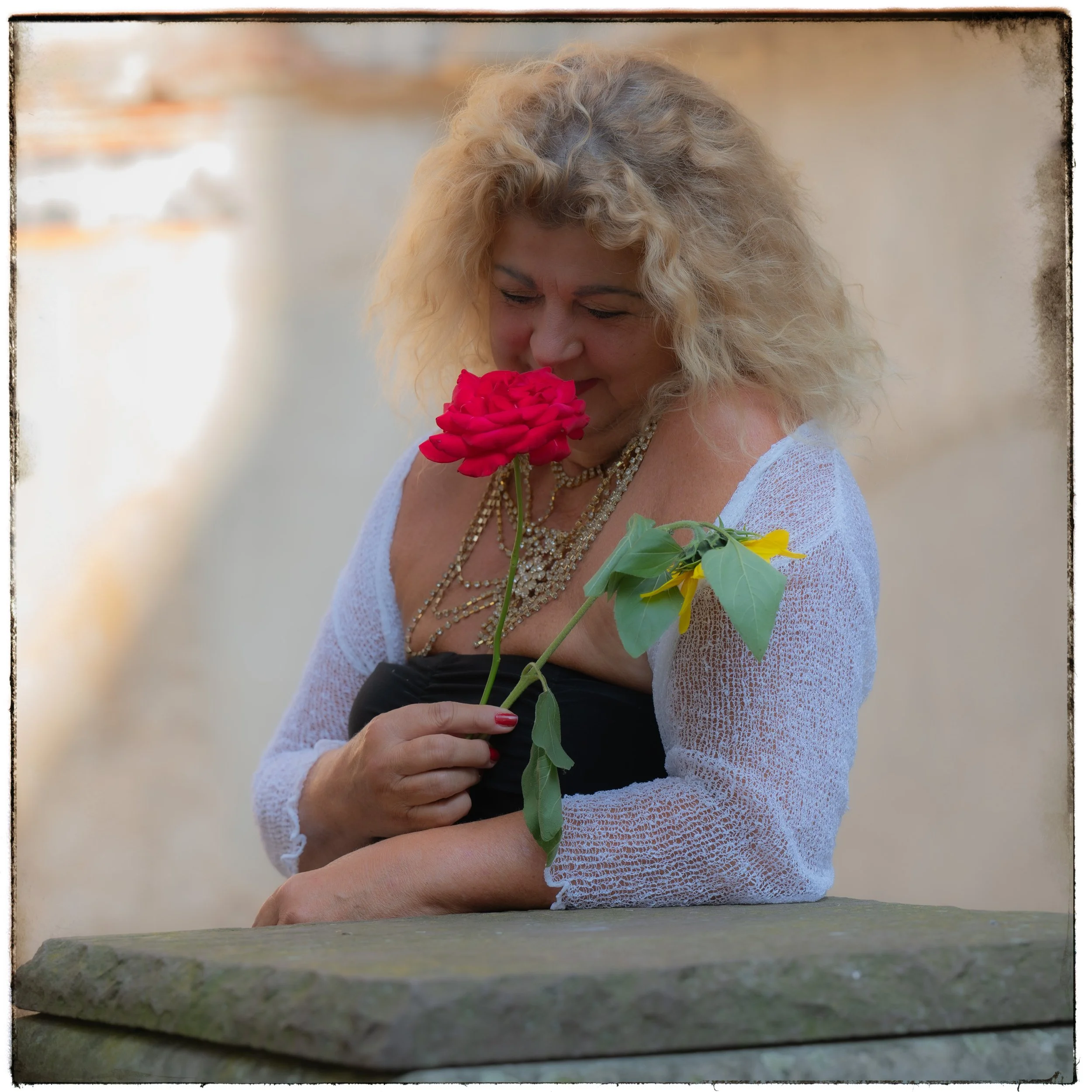 Eine blonde Frau, die eine rote Rose und eine gelbe Blume hält, lächelnd und die Augen geschlossen.