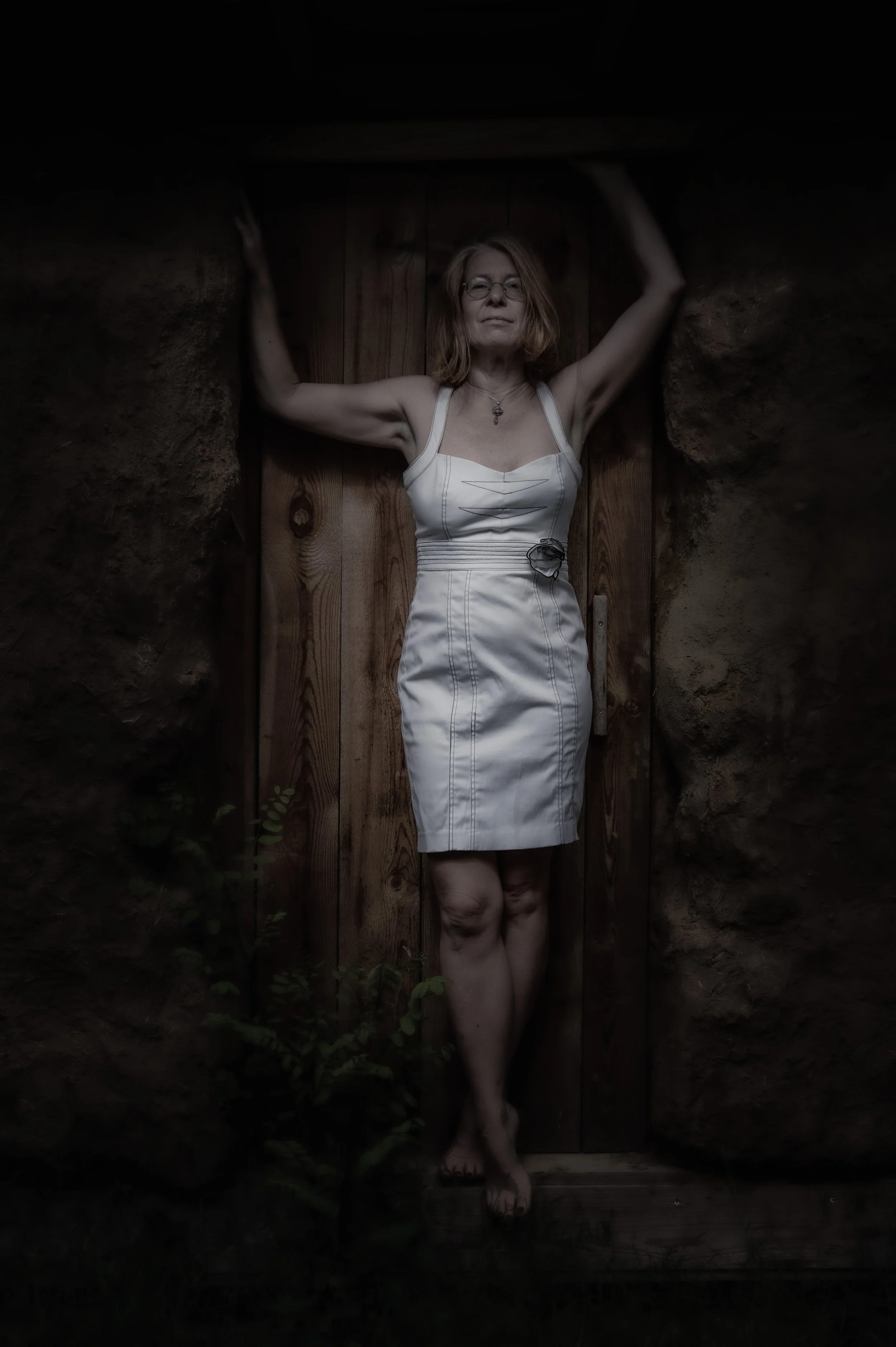 Eine Frau in einem weißen Kleid steht mit ausgestreckten Armen zwischen Holztür und Felswand, nackt bis auf die Beine, bei Dämmerlicht.