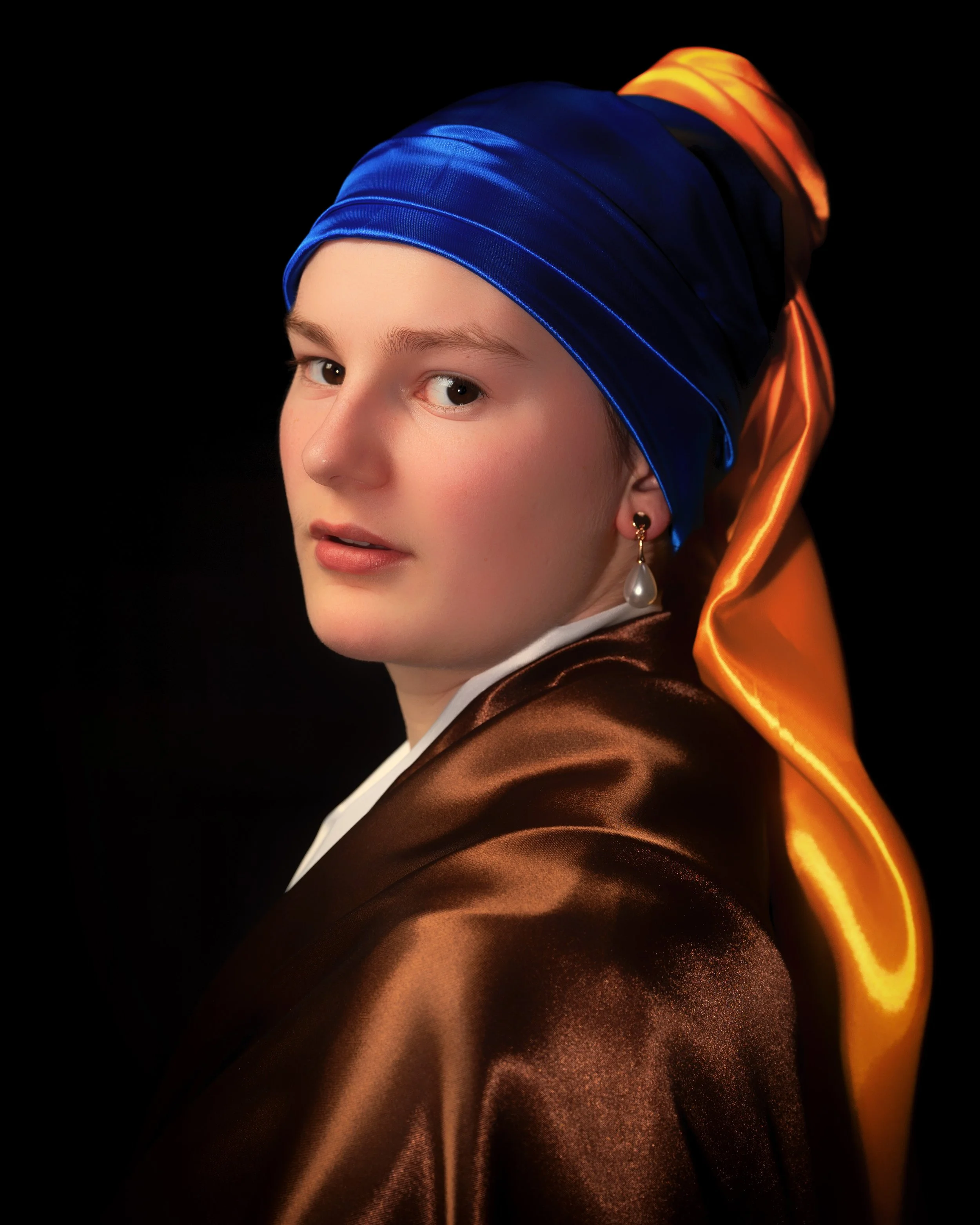 Porträt eines Mädchens in traditionellen Kleidung mit blauer und goldener Kopfbedeckung und braunem Mantel vor schwarzem Hintergrund.