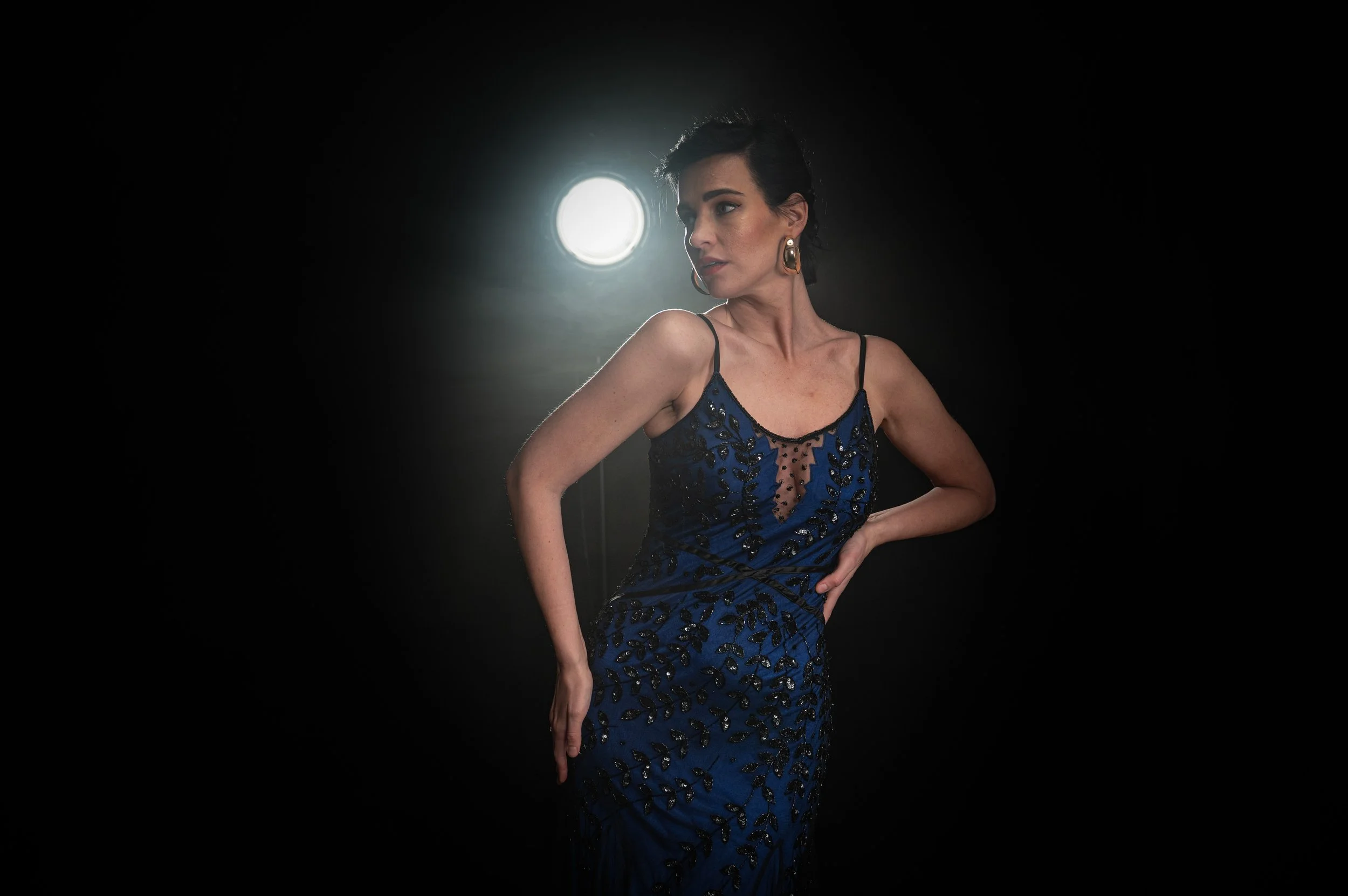 Frau in einem eleganten blauen Kleid mit schwarzen Mustern vor dunklem Hintergrund, mit Lichtquelle hinter ihr.