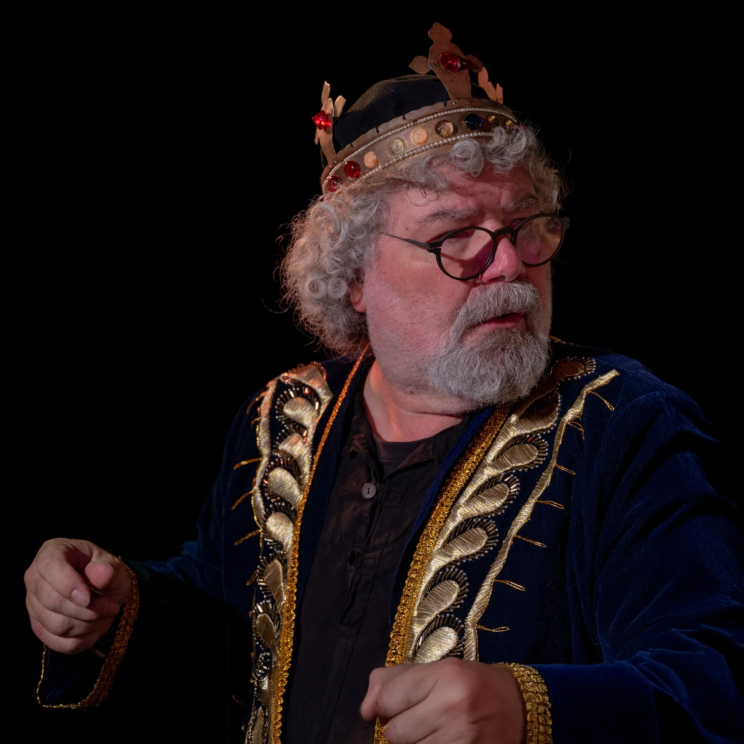 Ein älterer Mann mit Brille, grauem lockigem Haar und Bart trägt eine königliche Krone und eine schwarze Jacke mit goldverzierten Stickereien, sitzt gegen einen schwarzen Hintergrund.