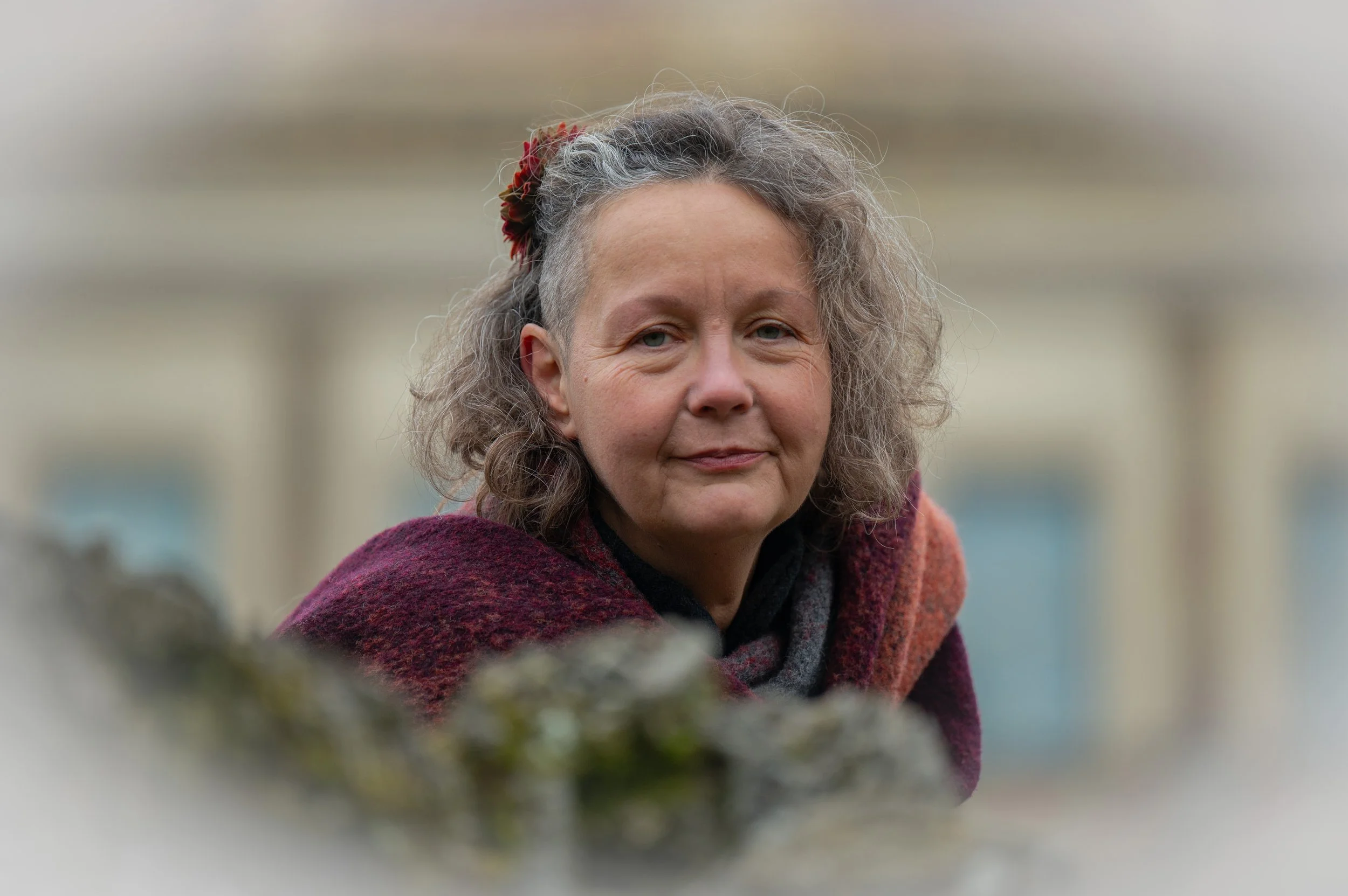 Porträt einer älteren Frau mit grauem, lockigem Haar, die einen roten Haarkamm trägt, vor einem unscharfen Hintergrund.