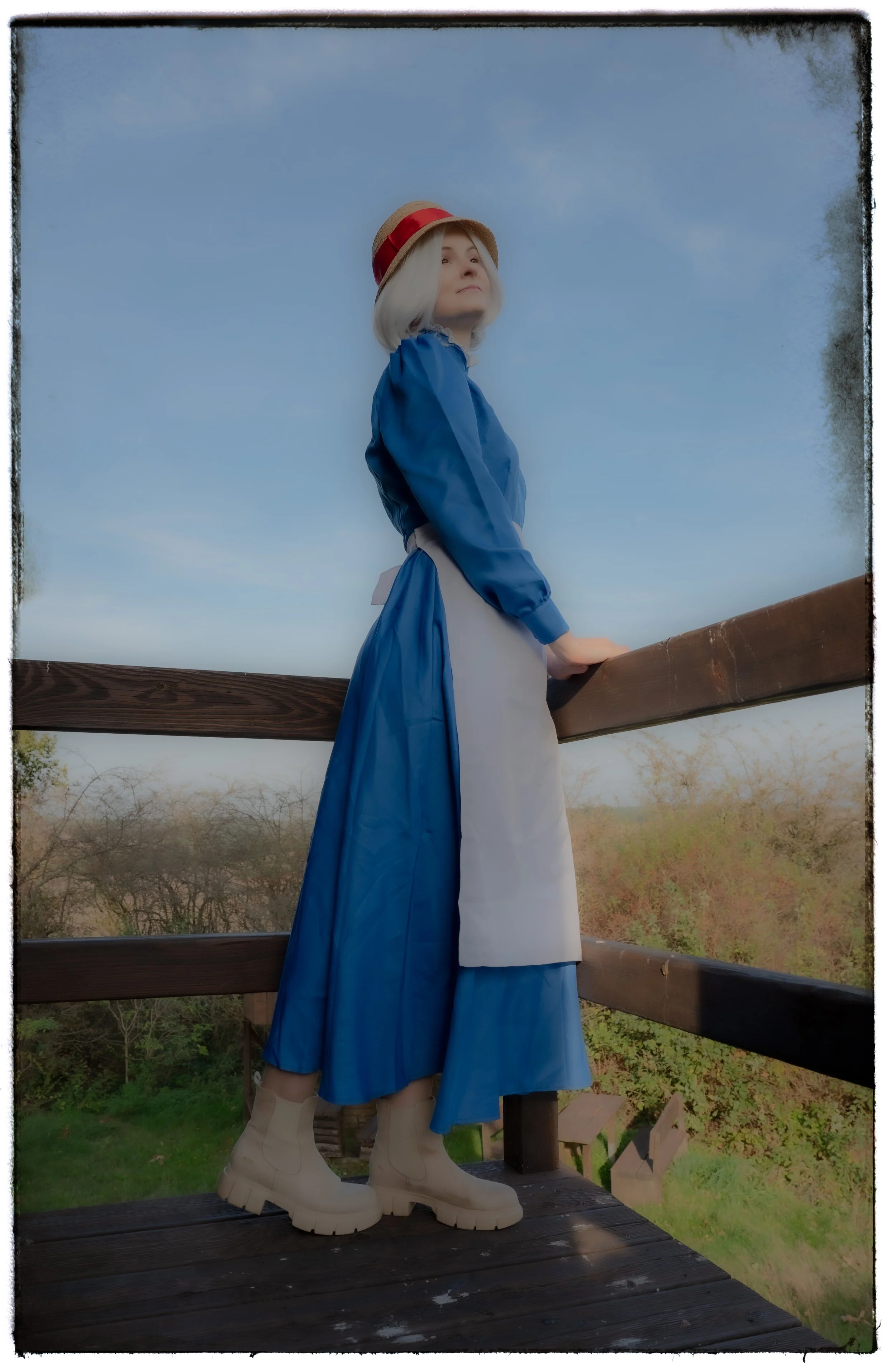 Frau in blauer Kleid mit Hut steht auf einer Holzplattform, Blick in die Ferne, im Hintergrund Bäume und Himmel.