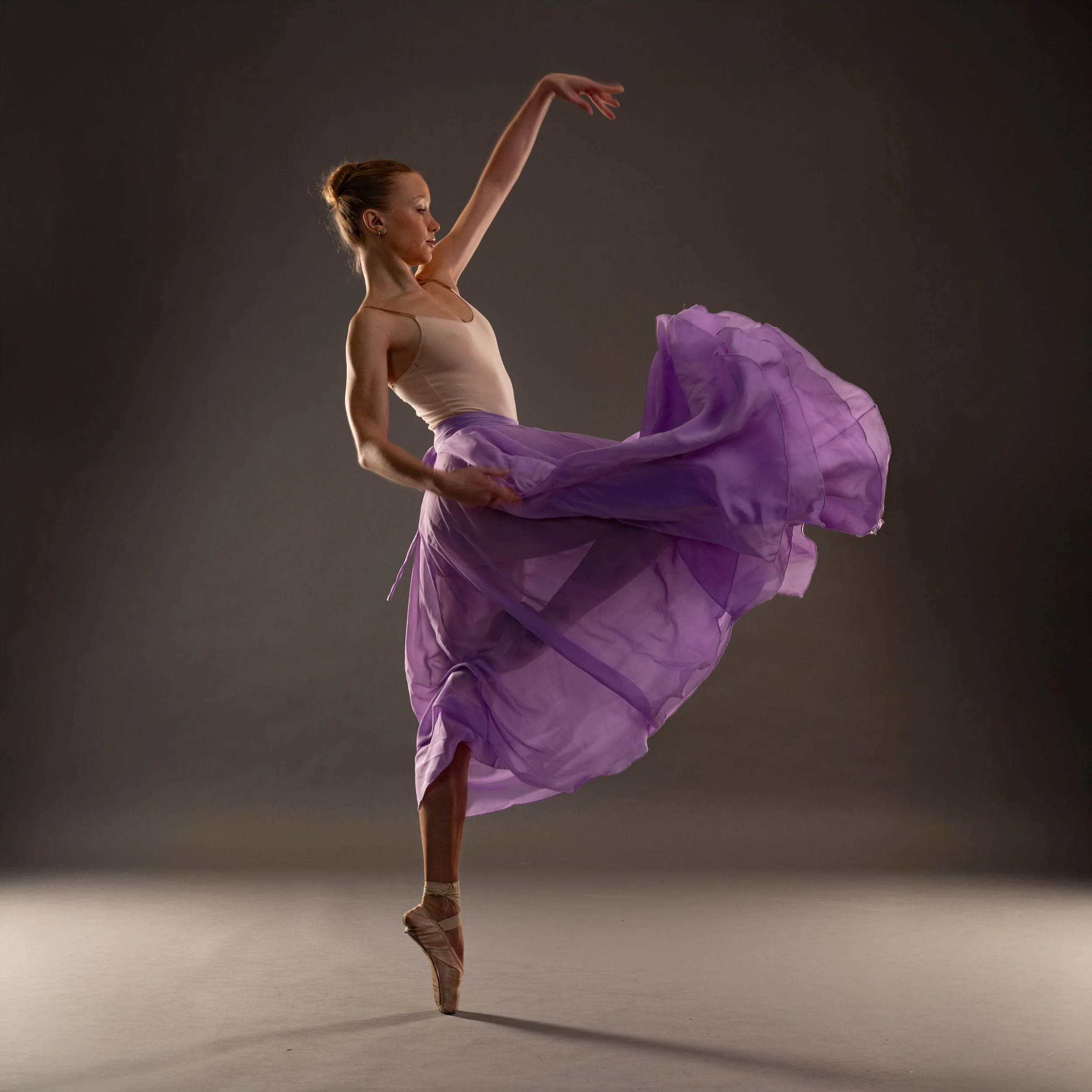 Balletttänzerin in pinkem Tutu und Ballettschuhen, die auf Spitze tanzt, vor grau-schattigem Hintergrund.