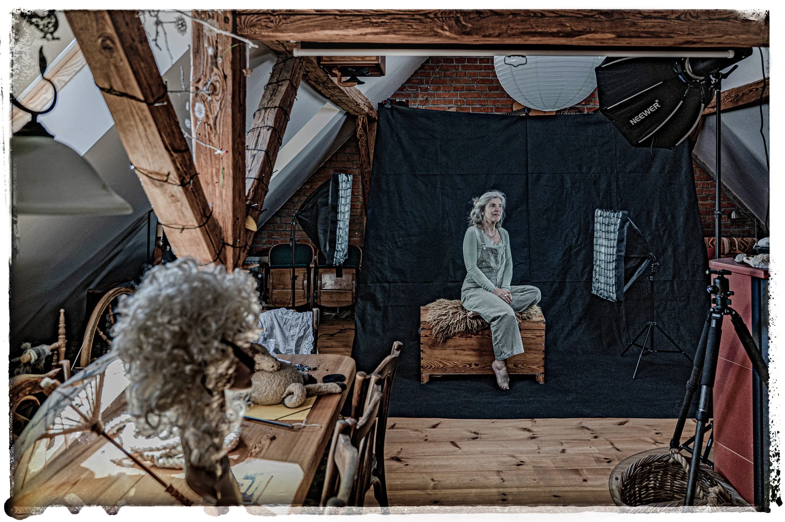 Eine Frau sitzt auf einer Holzbox vor einem schwarzen Hintergrund in einem Raum mit sichtbaren Dachbalken und kreativer Inneneinrichtung. Es sieht nach einem Fotostudio aus.