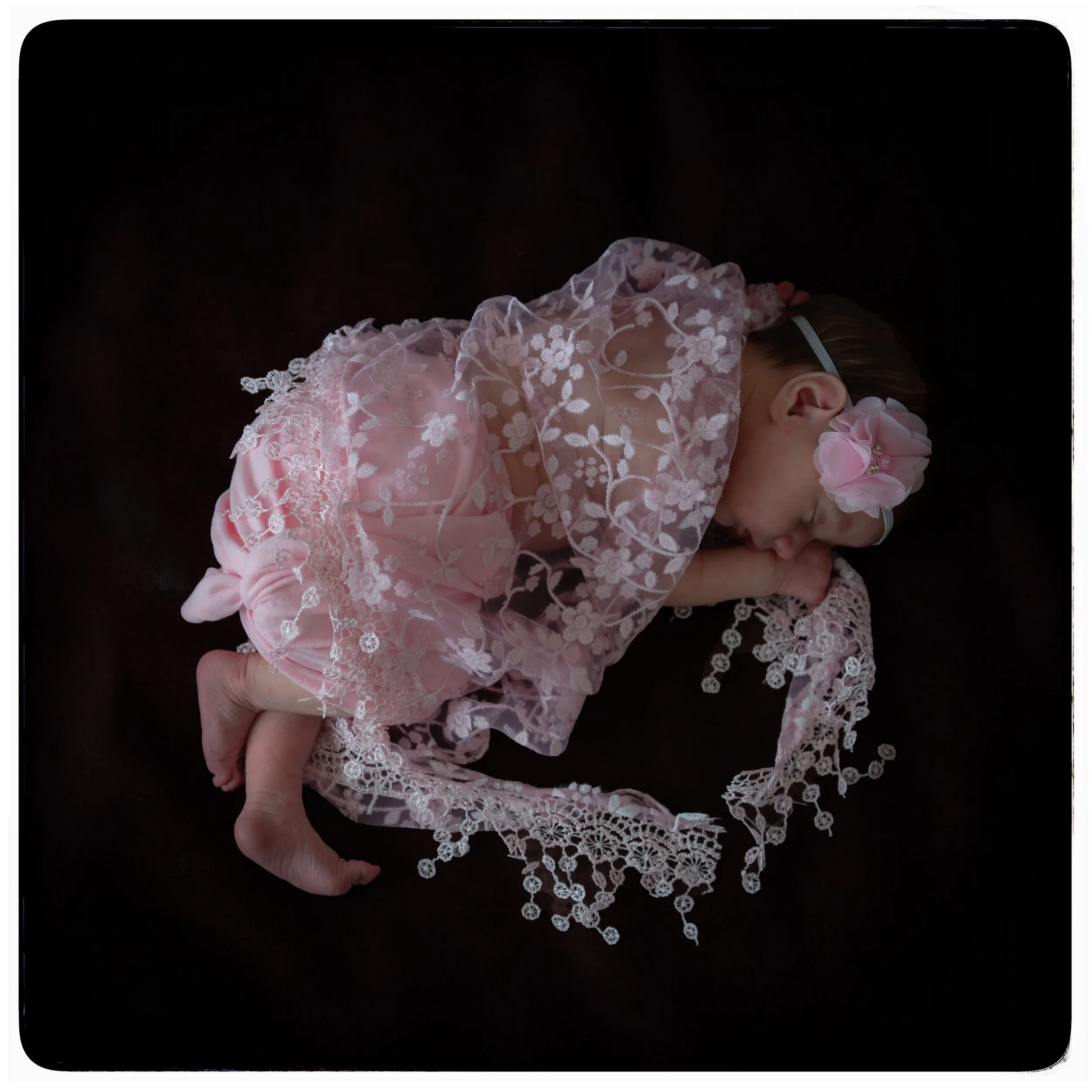 Kleines Baby, eingekleidet in rosene Kleidung mit Spitzenkragen, liegt schlafend auf einem dunklen Hintergrund. Das Baby trägt eine große rosa Blume im Haar.