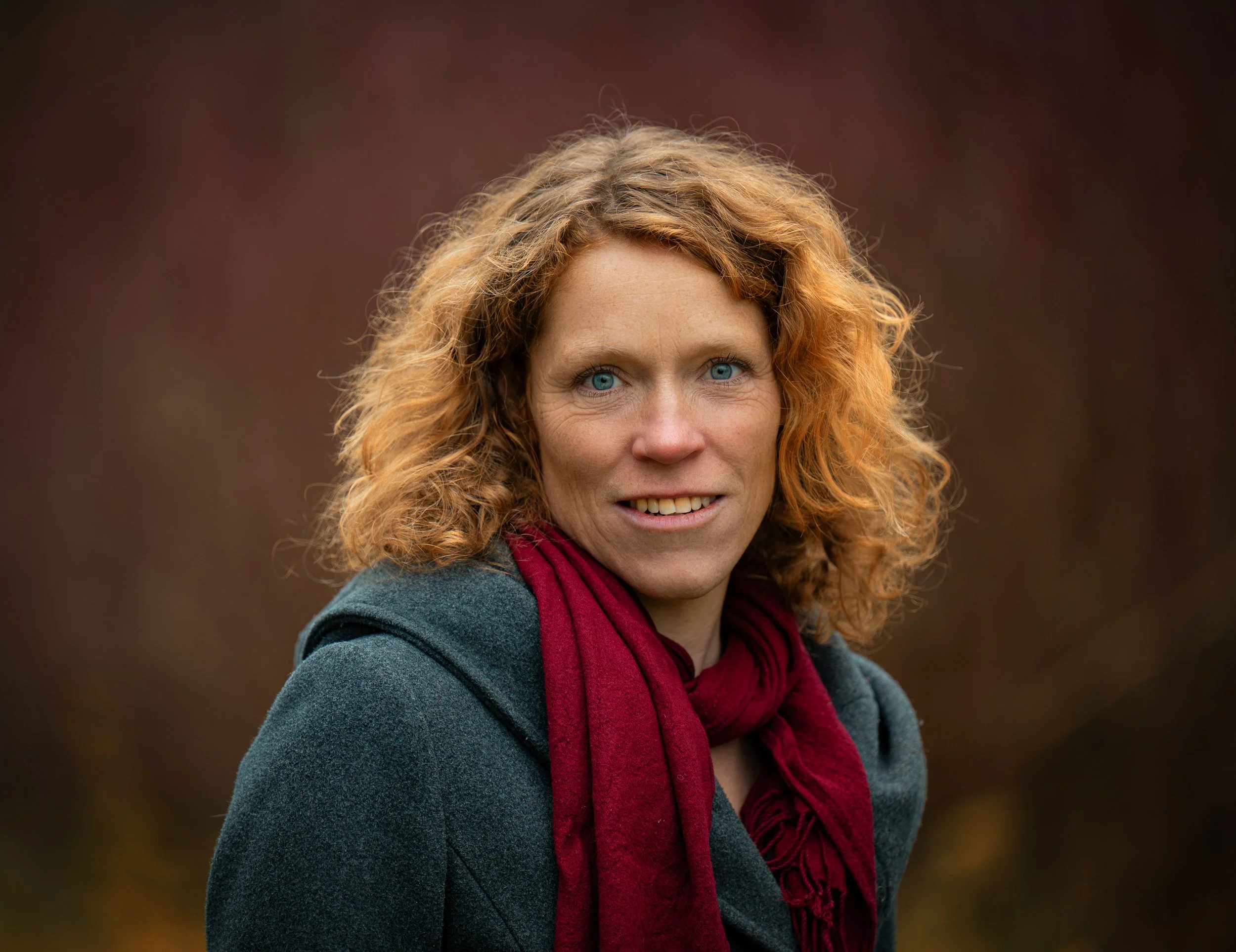 Frau mit roten, lockigen Haaren, blaue Augen, grauem Mantel und roter Schal, im Freien bei bewölktem Himmel.