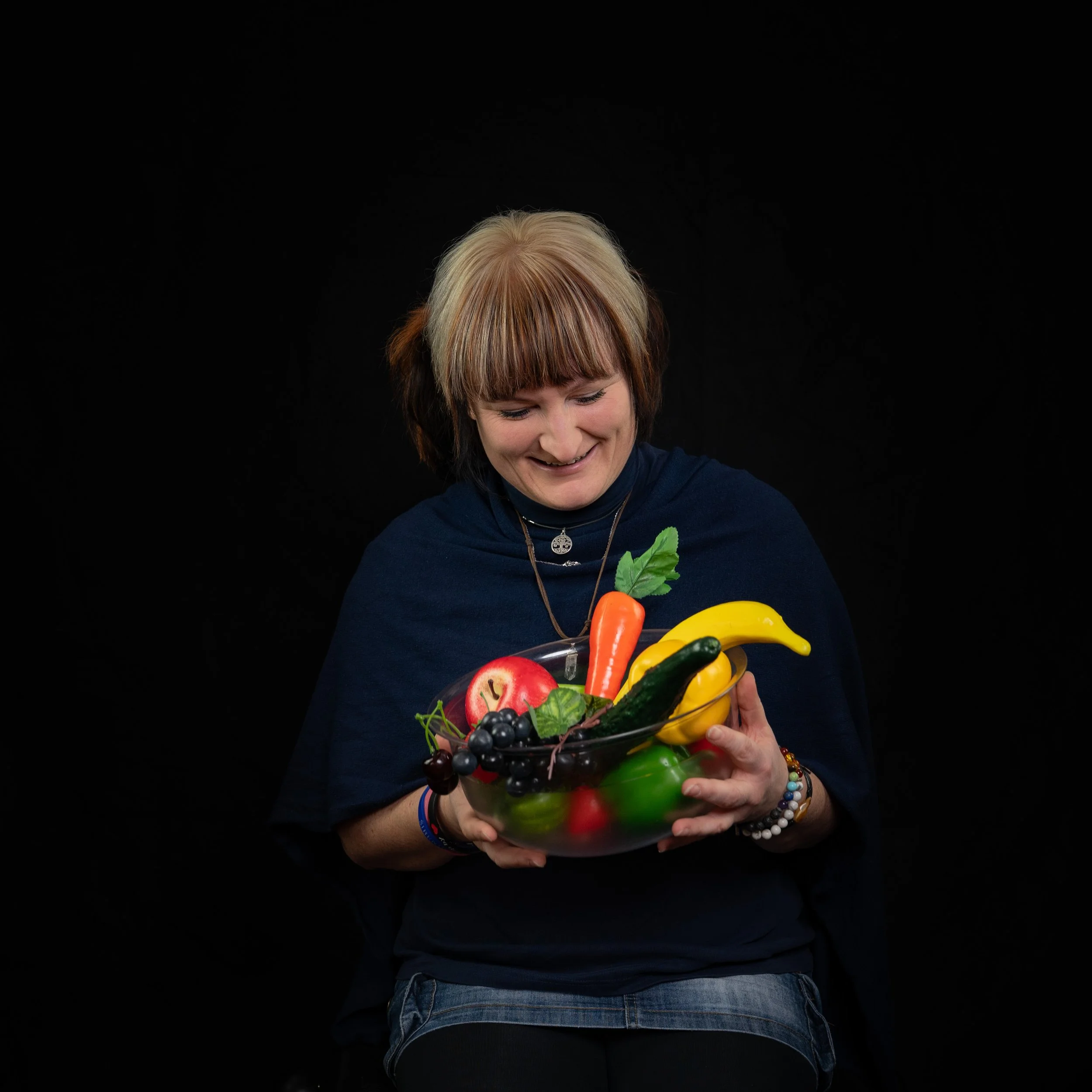 Eine Frau hält eine Schüssel mit buntem Obst und Gemüse, darunter Äpfel, Trauben, eine Karotte, Gurke, Paprika und Bananen.