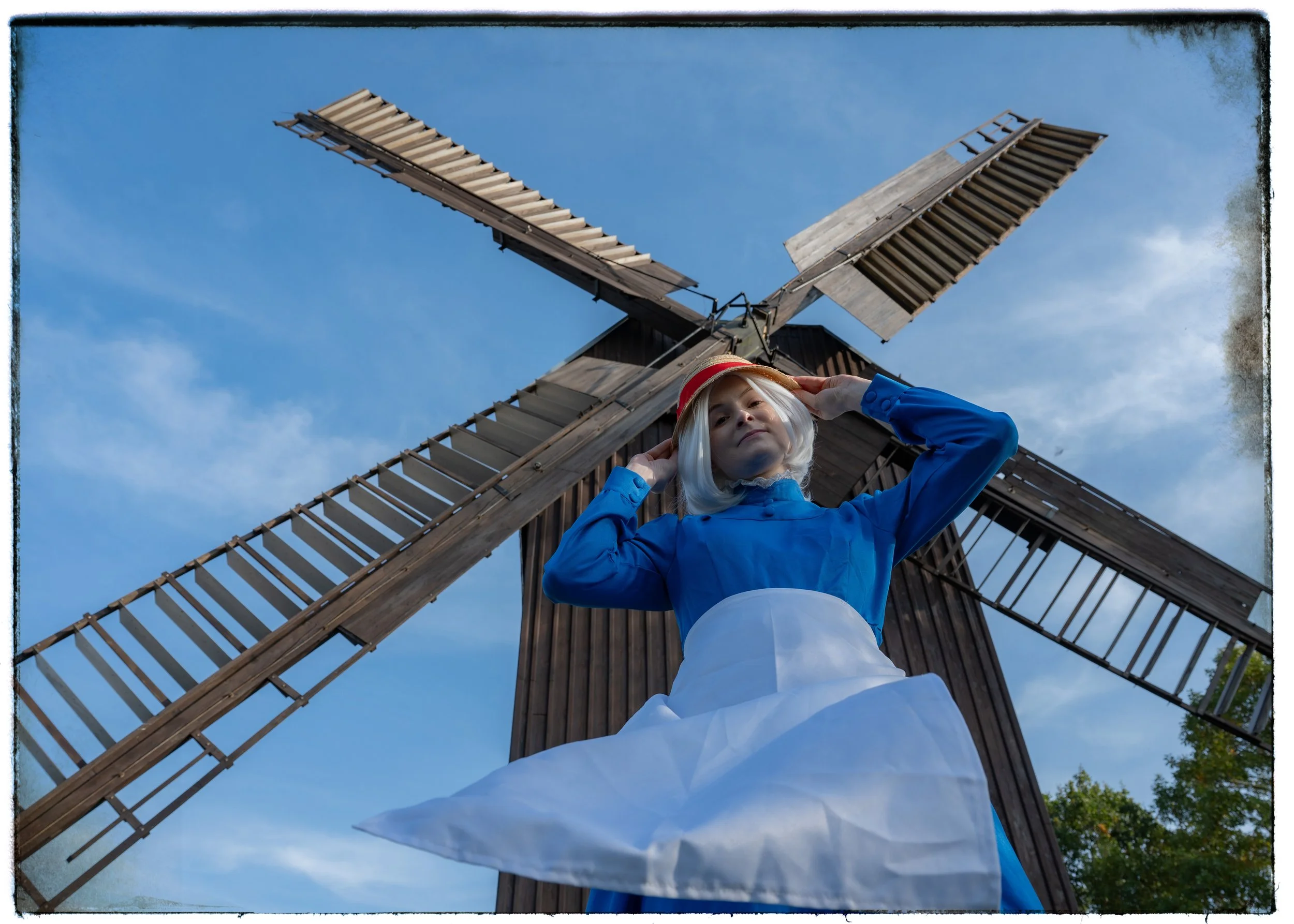 Puppe im viktorianischen Kleid und mit Hut, steht vor einem alten Mühlengebäude, aufgenommen von unten, gegen einen blauen Himmel