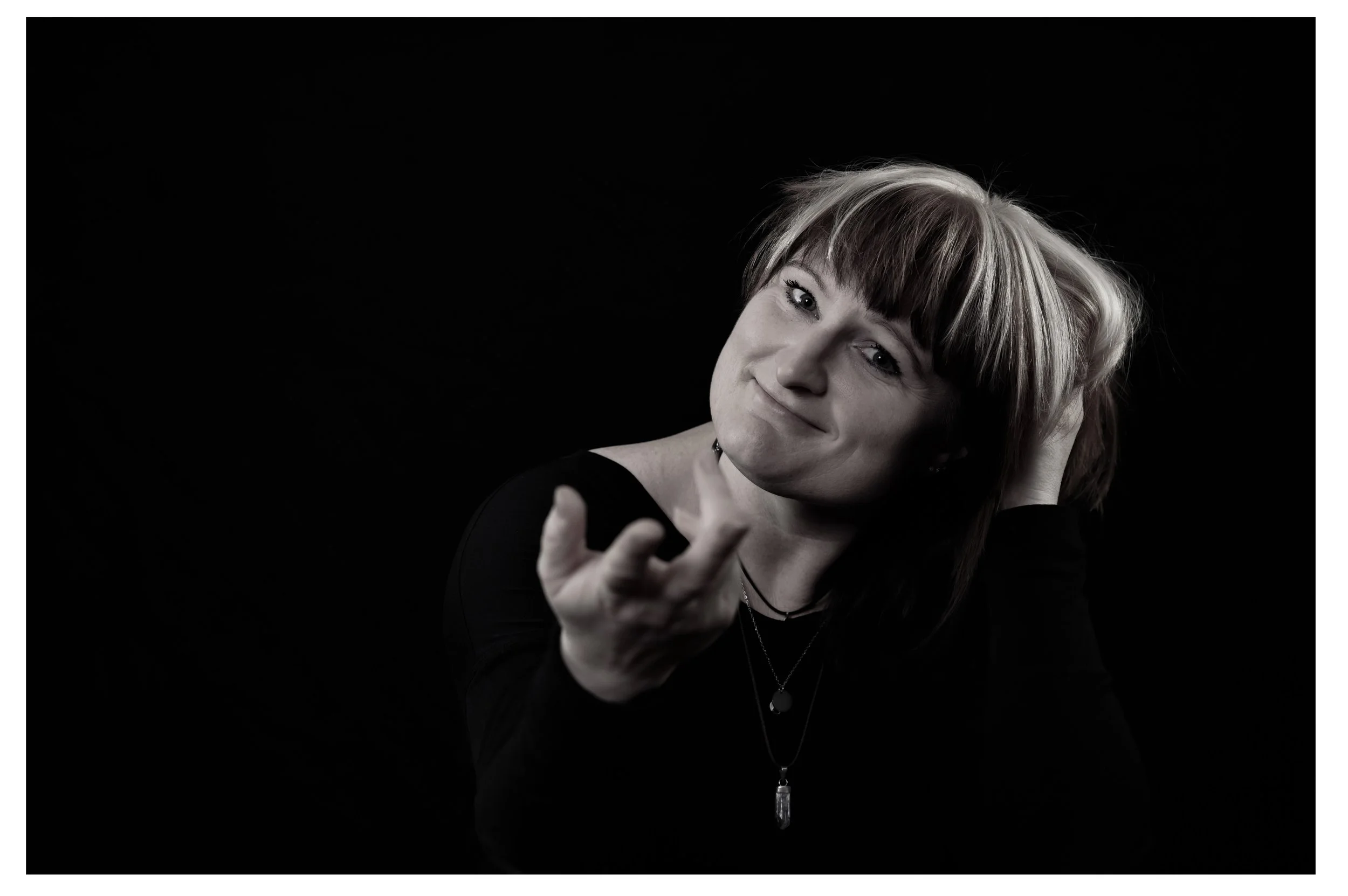 Junge Frau mit Kurzhaarfrisur und dunklem Outfit, die in die Kamera lächelnd ihre Hand ausstreckt, vor schwarzem Hintergrund.