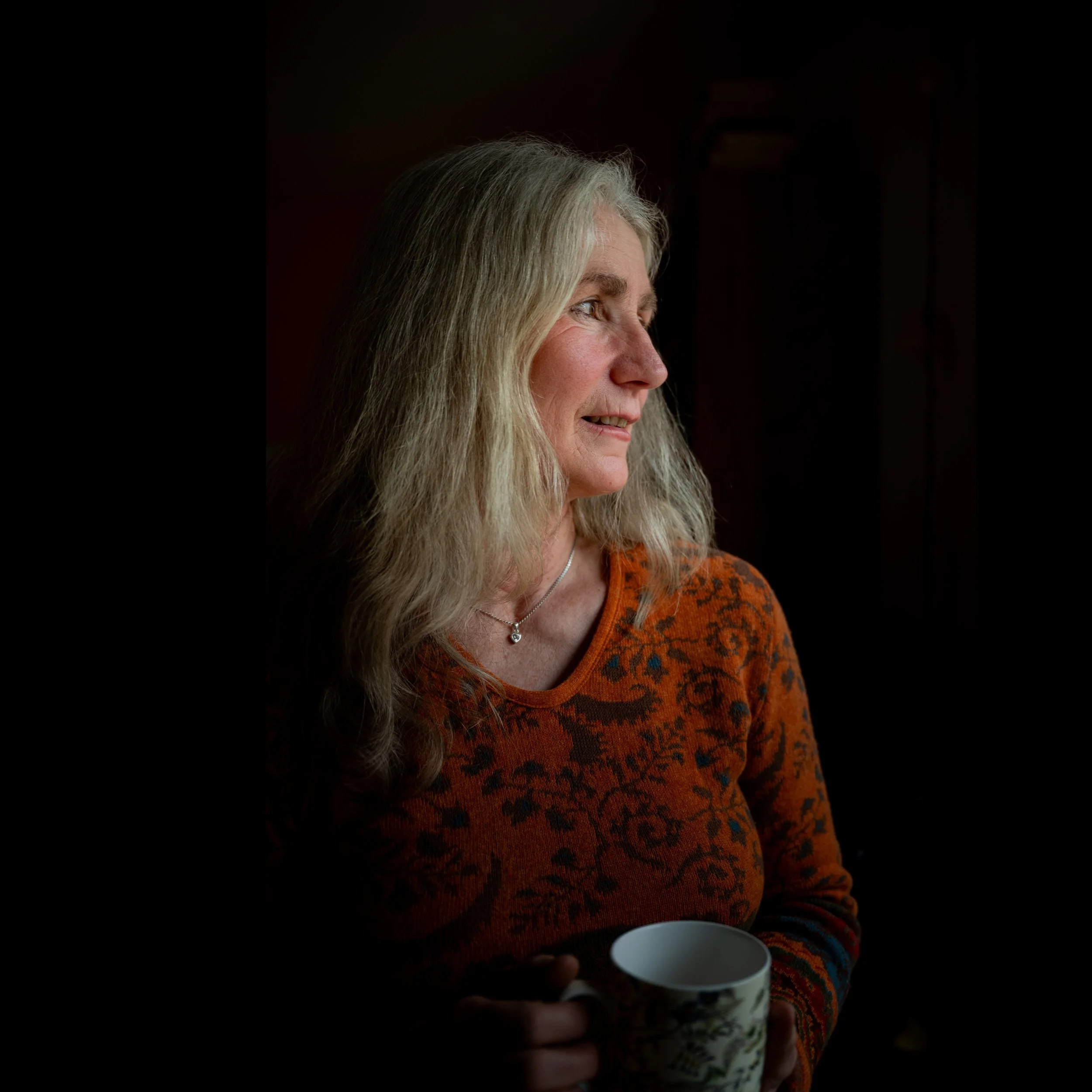 Eine ältere blonde Frau trägt ein orangefarbenes, gemustertes Oberteil und hält eine Tasse. Sie blickt nach rechts, im Hintergrund ist ein dunkler Raum.