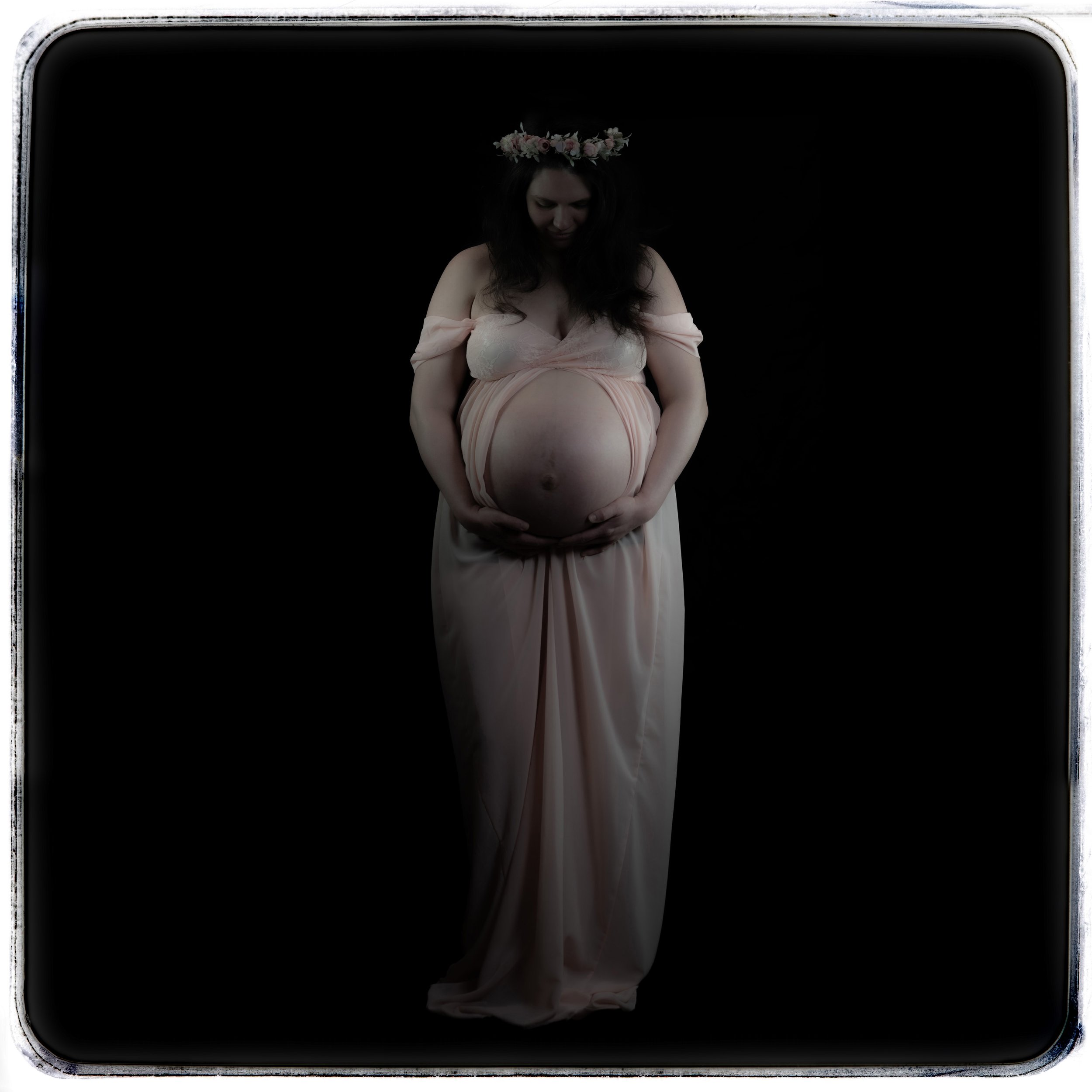 Schwangere Frau mit Blumenkrone im hellen Kleid vor schwarzem Hintergrund.