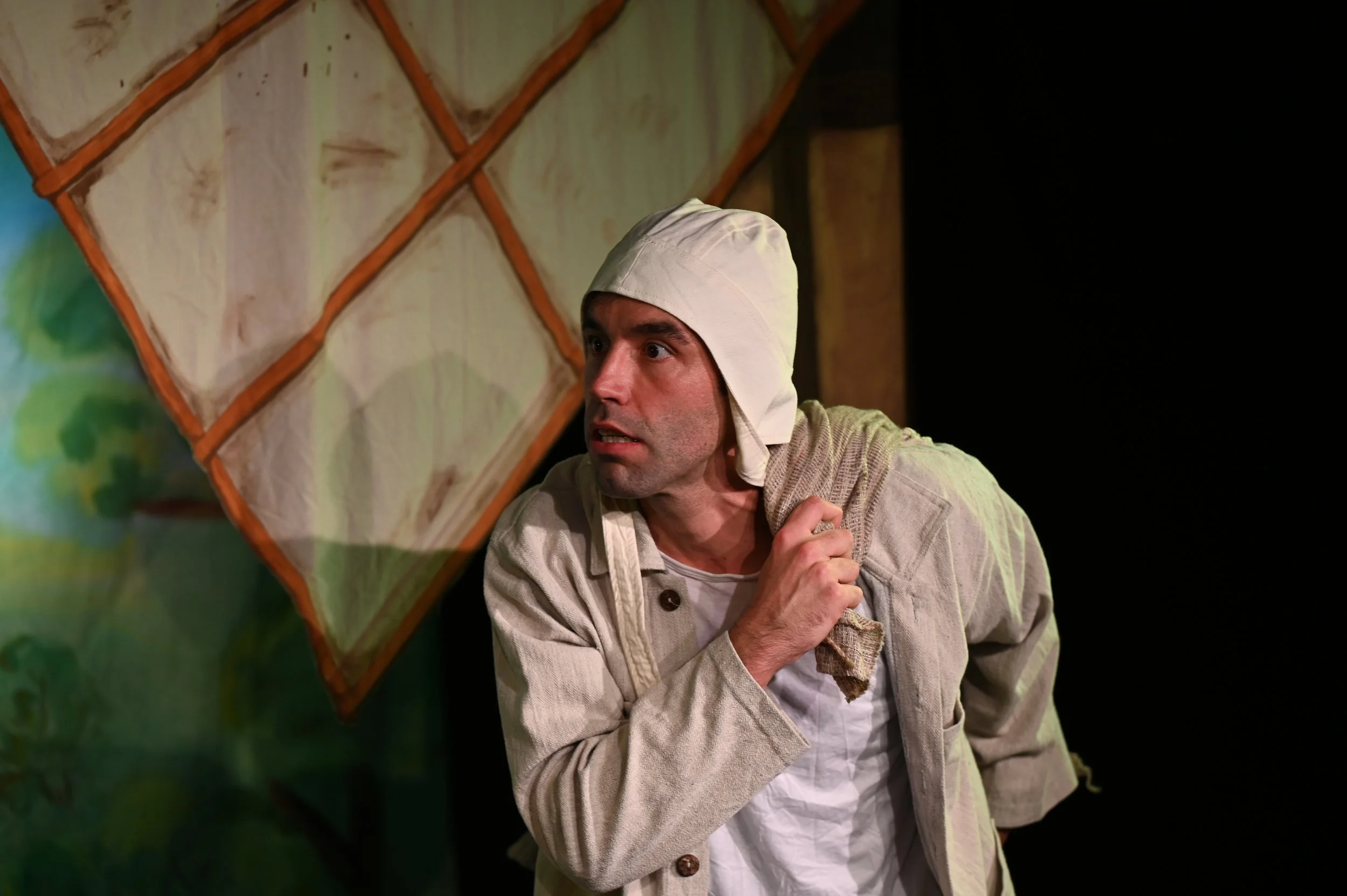 Ein Mann trägt ein weißes Tuch auf dem Kopf, sieht ängstlich aus und croucht hinter einer Wand auf einer Bühne.