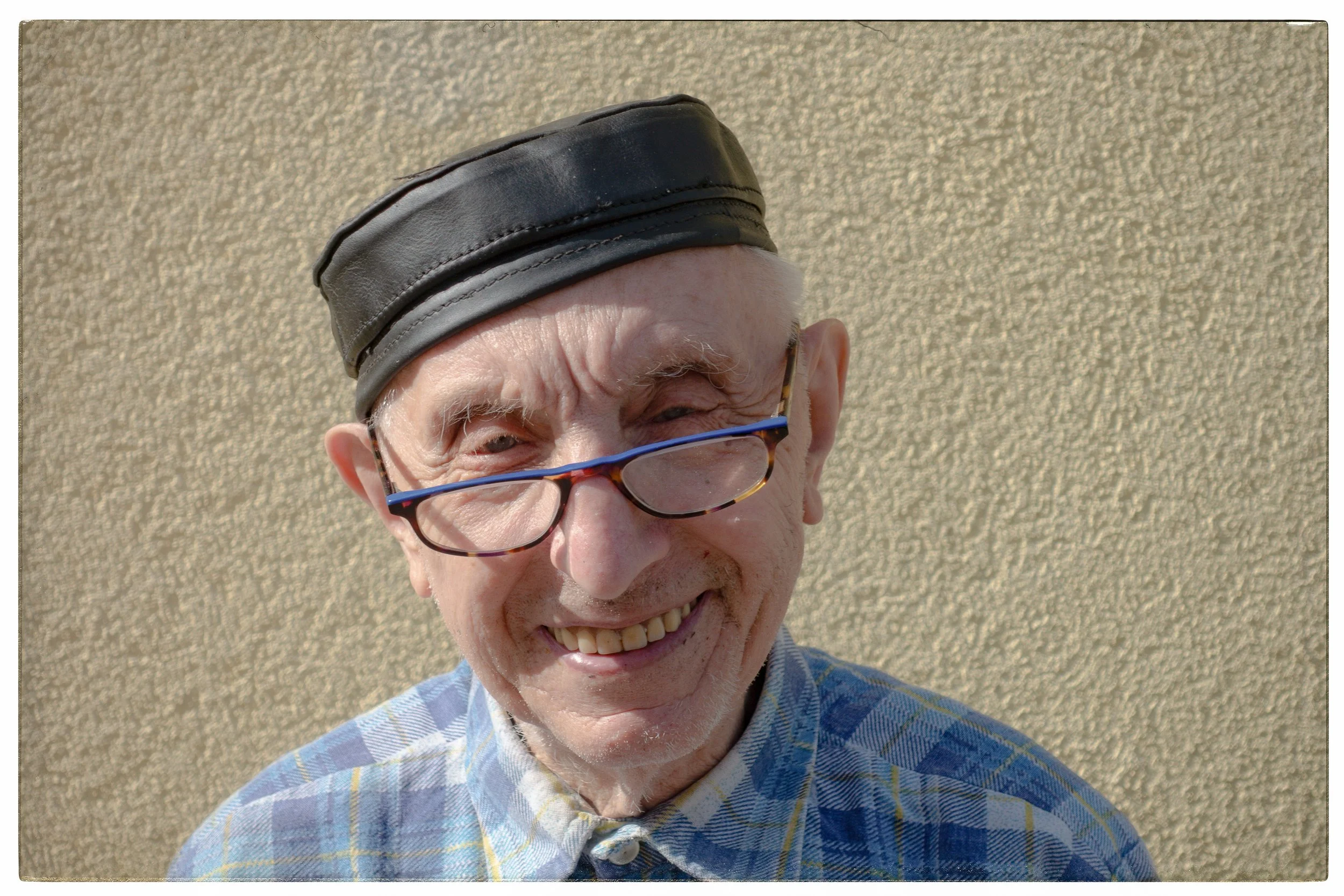 Älterer Mann mit Brille und schwarzem Lederhut, lächelt vor einer beige Wand.