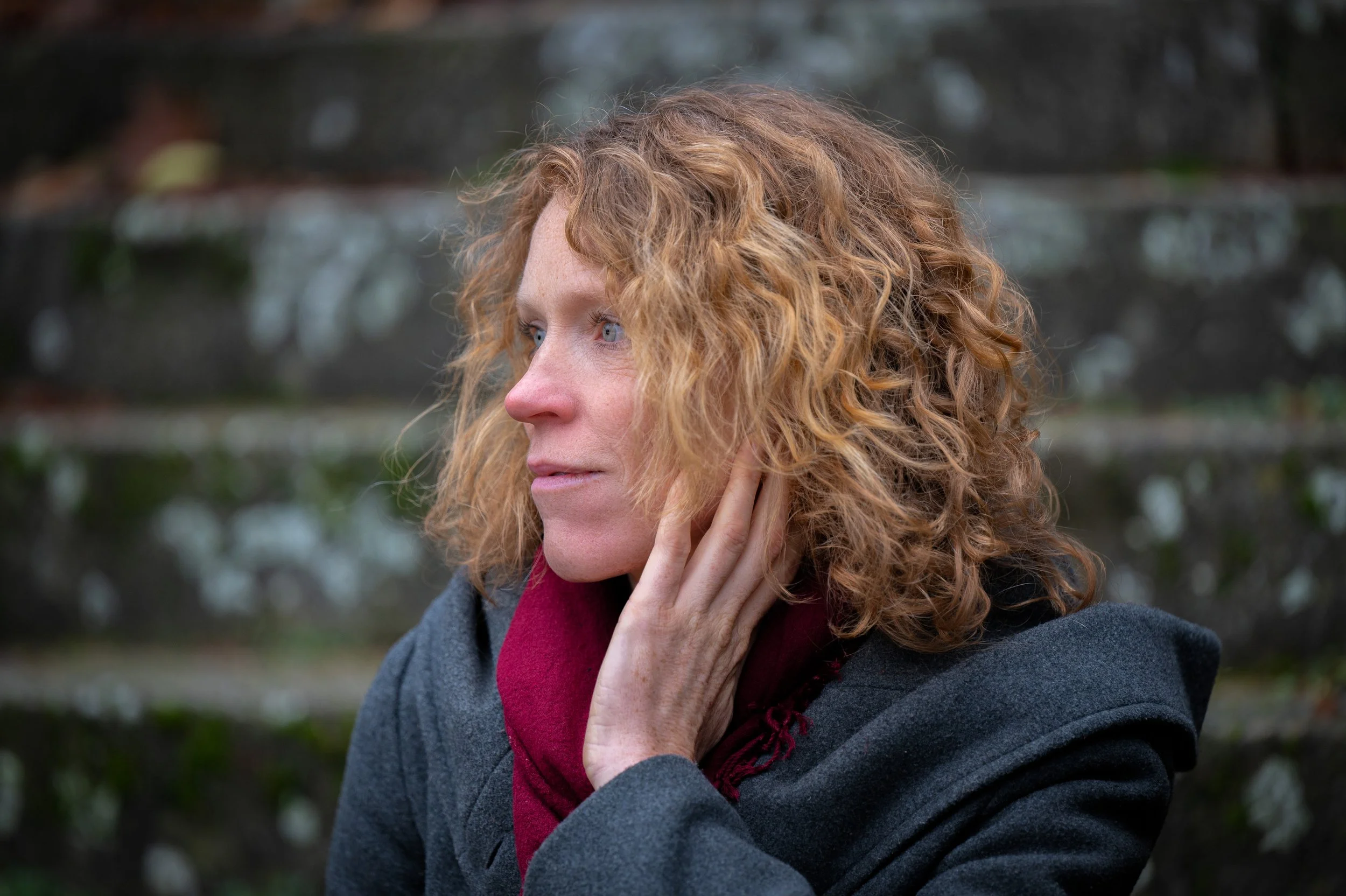 Nahaufnahme einer Frau mit lockigem roten Haar, die nach links schaut, die Hand an ihrem Hals hält und vor einem Steingarten steht.