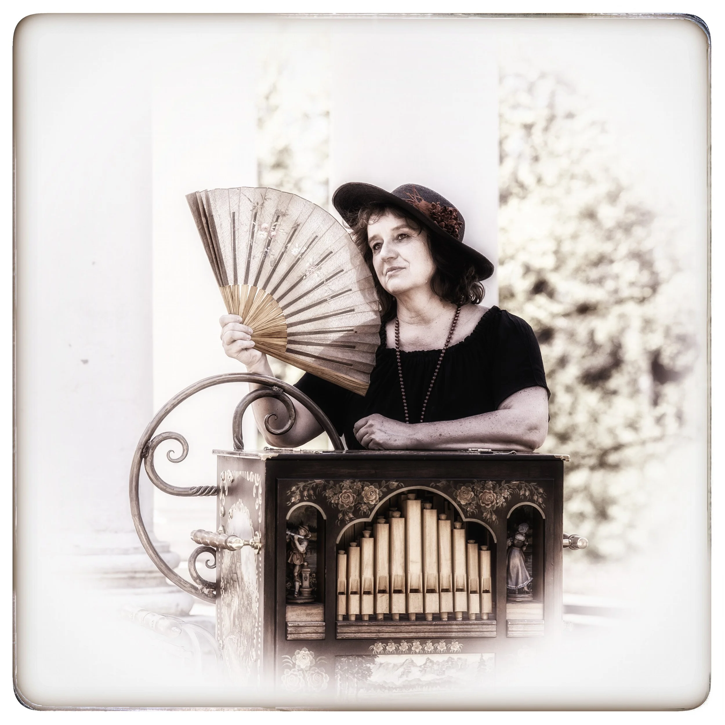 Eine ältere Frau mit dunklem Hut und schwarzem Kleid, die ein Fächer hält, steht neben einem historischen Orgelkasten im Freien.