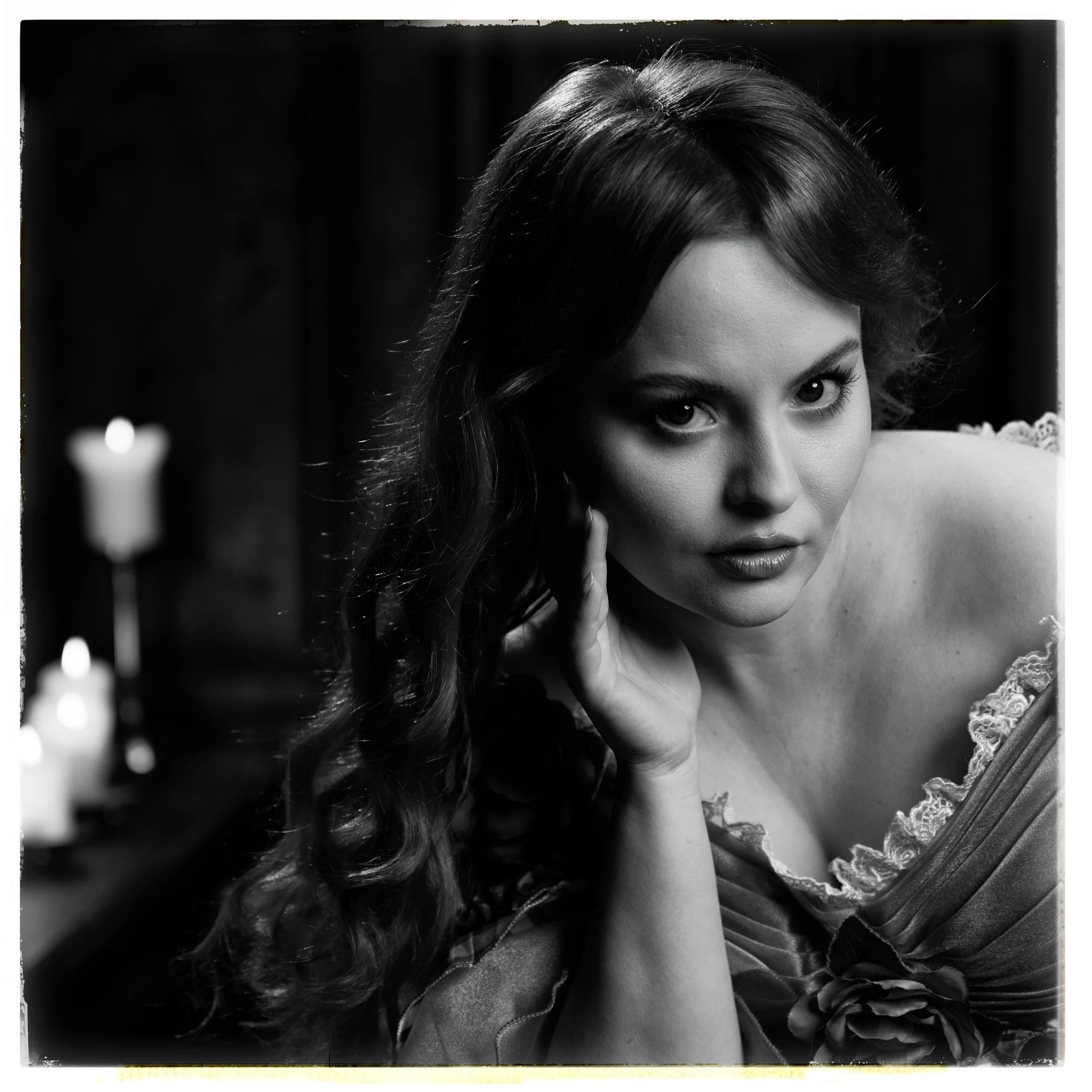 Schwarz-weiß Foto einer jungen Frau mit lockigem Haar, die direkt in die Kamera schaut, die Hand an ihrem Gesicht hält und ein Kleid mit Lace-Details trägt, im Hintergrund sind Kerzen sichtbar.