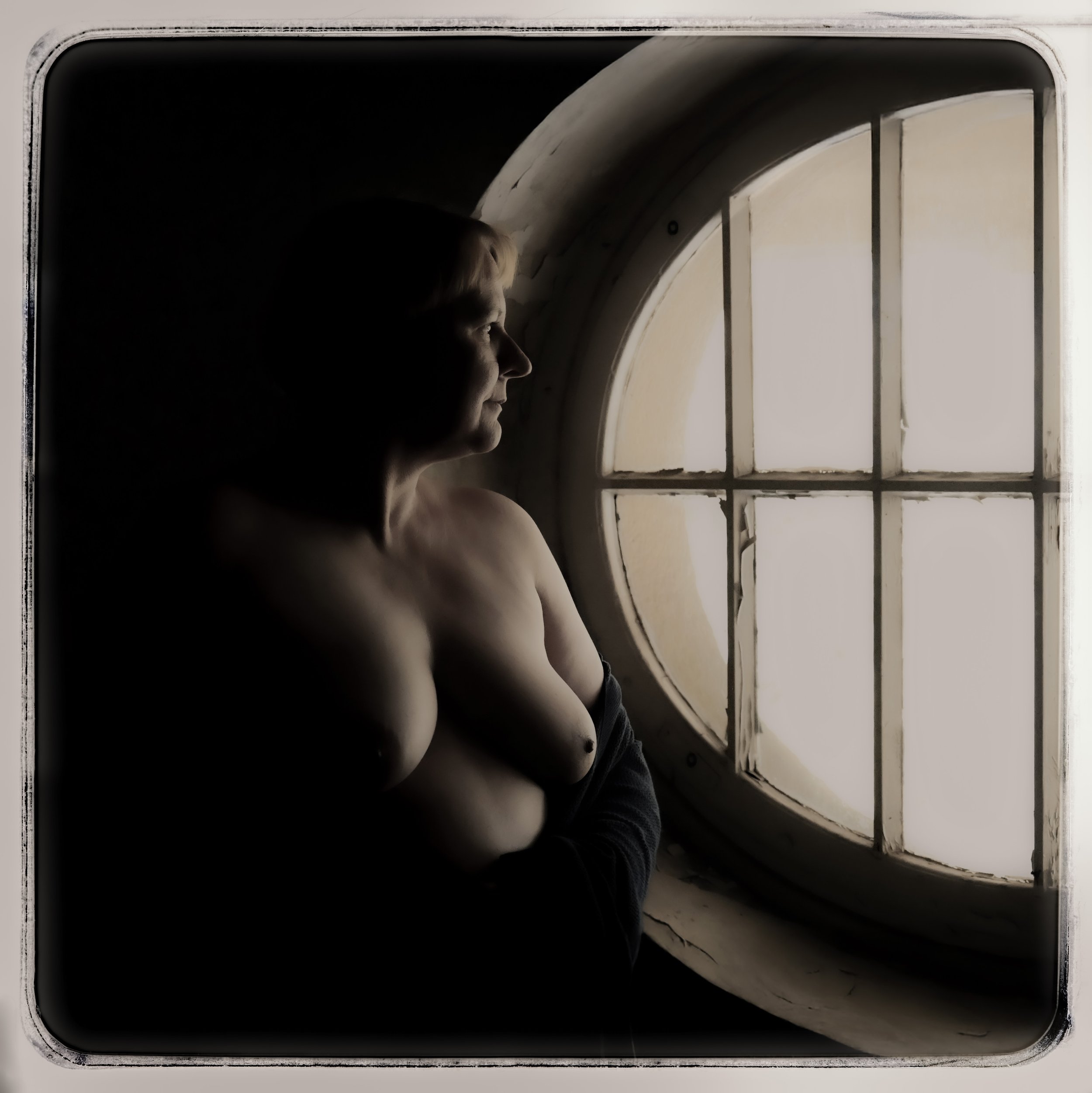 Eine nackte Frau steht am Fenster und schaut nach draußen, die Szene ist im Schatten gehalten, mit Licht vom Fenster beleuchtet.