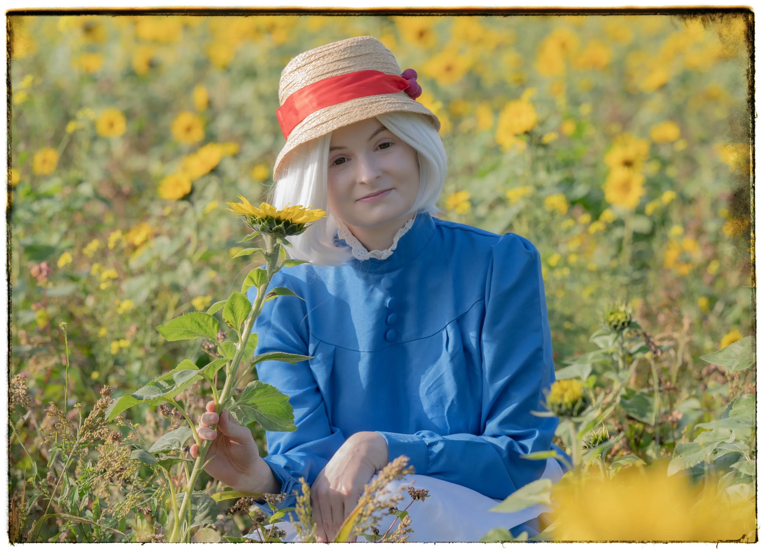 Eine junge Frau mit weißem Haar, blauem Kleid und Strohhut sitzt in einem Sonnenblumenfeld und hält eine große Sonnenblume in der Hand.