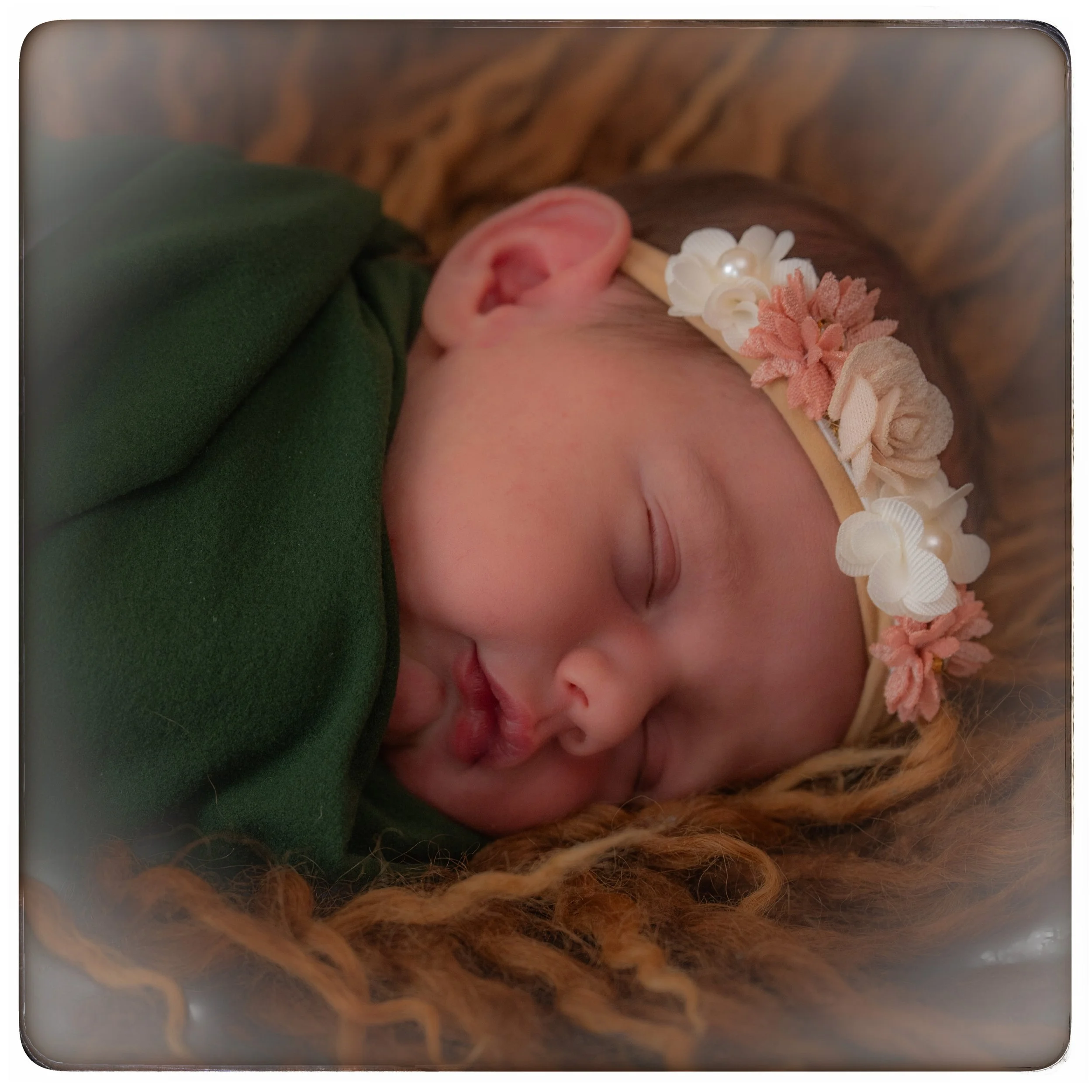 Ein schlafendes Baby mit einem festlichen Haarreif mit Blumen und Perlen, eingewickelt in dunkle grüne Kleidung, liegt auf einem braunen, geflochtenen Untergrund.