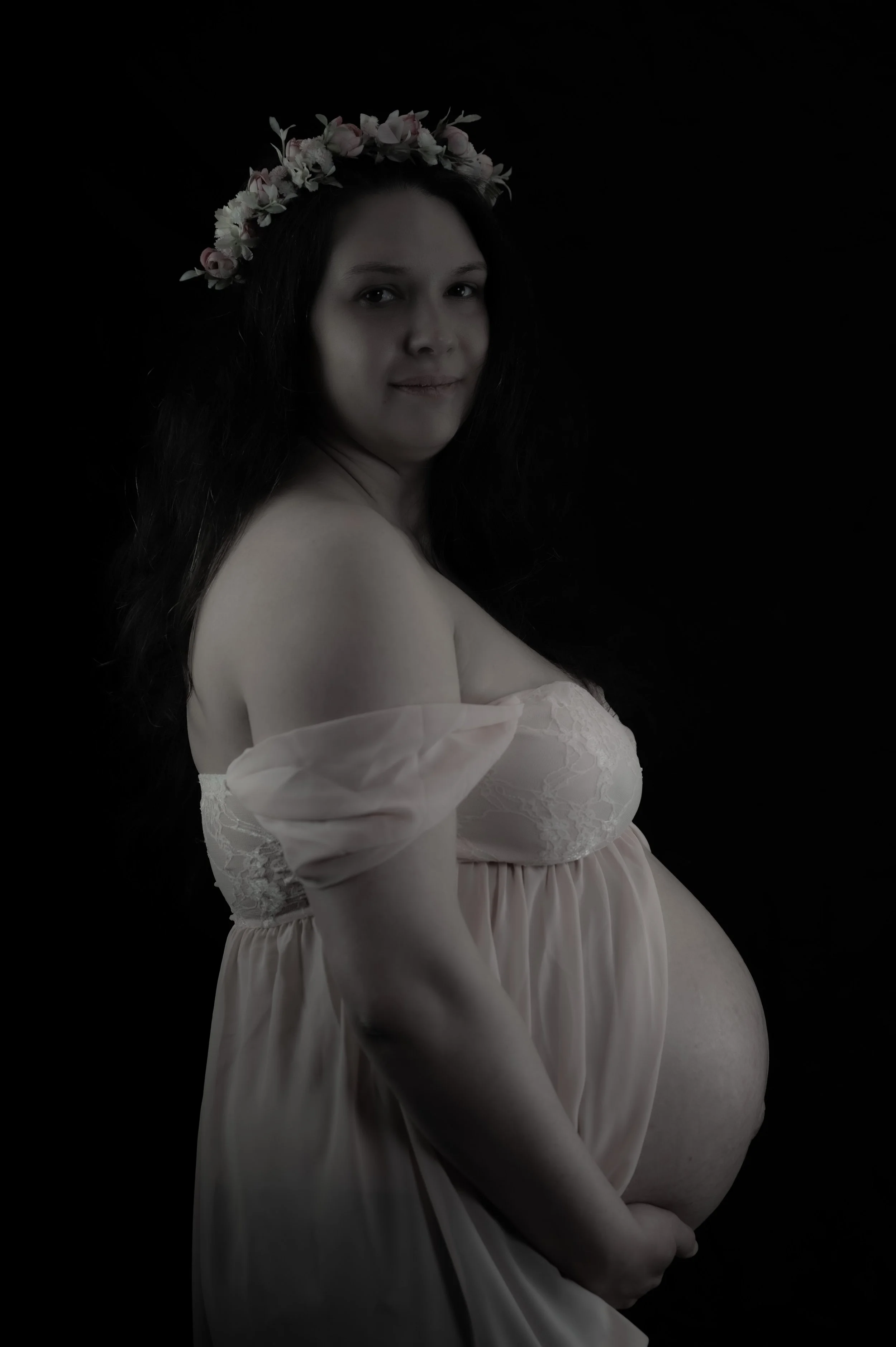 Schwangere Frau mit Blumenkranz auf dem Kopf trägt ein helles, fließendes Kleid gegen schwarzen Hintergrund.
