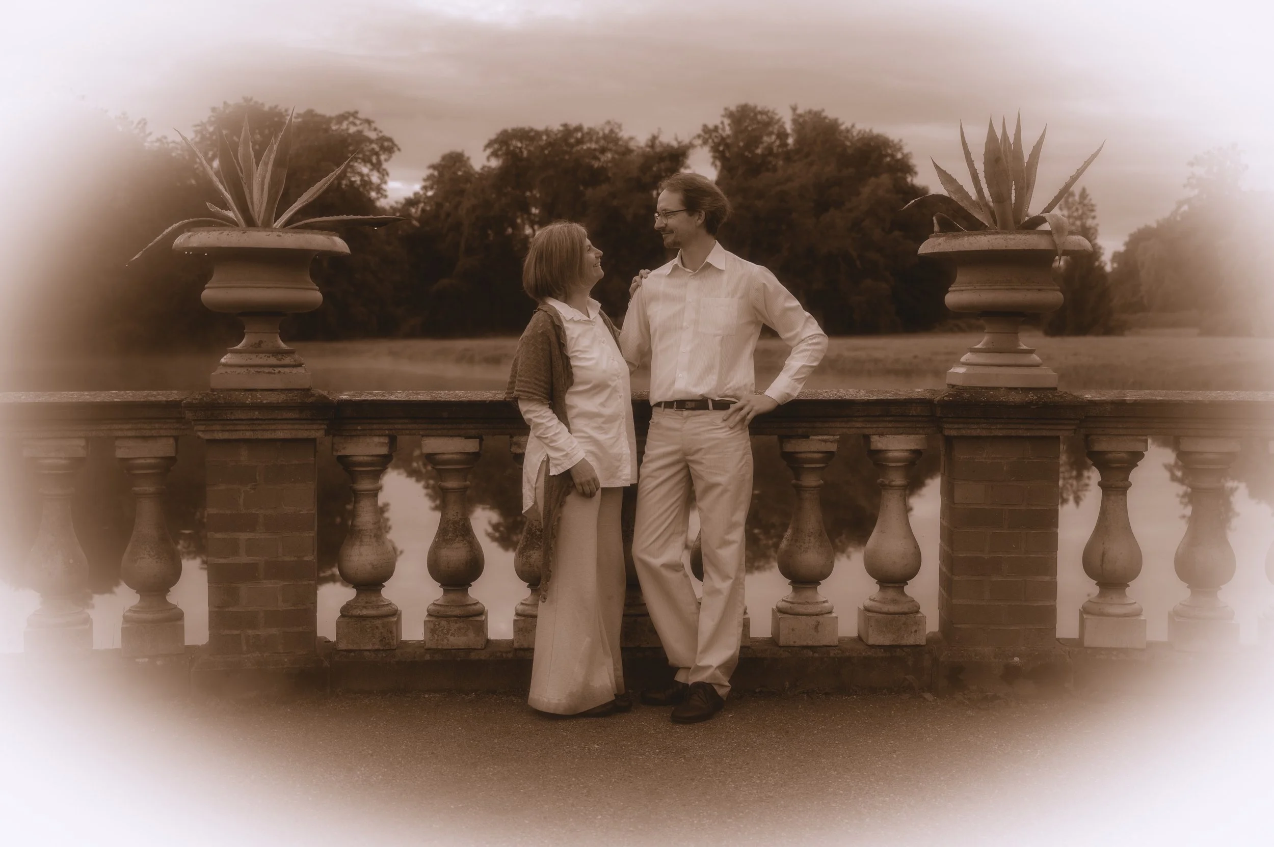 Ein älteres Paar steht auf einer Brüstung, umgeben von großen Topfpflanzen, und schaut sich liebevoll an, mit einer Naturkulisse im Hintergrund.