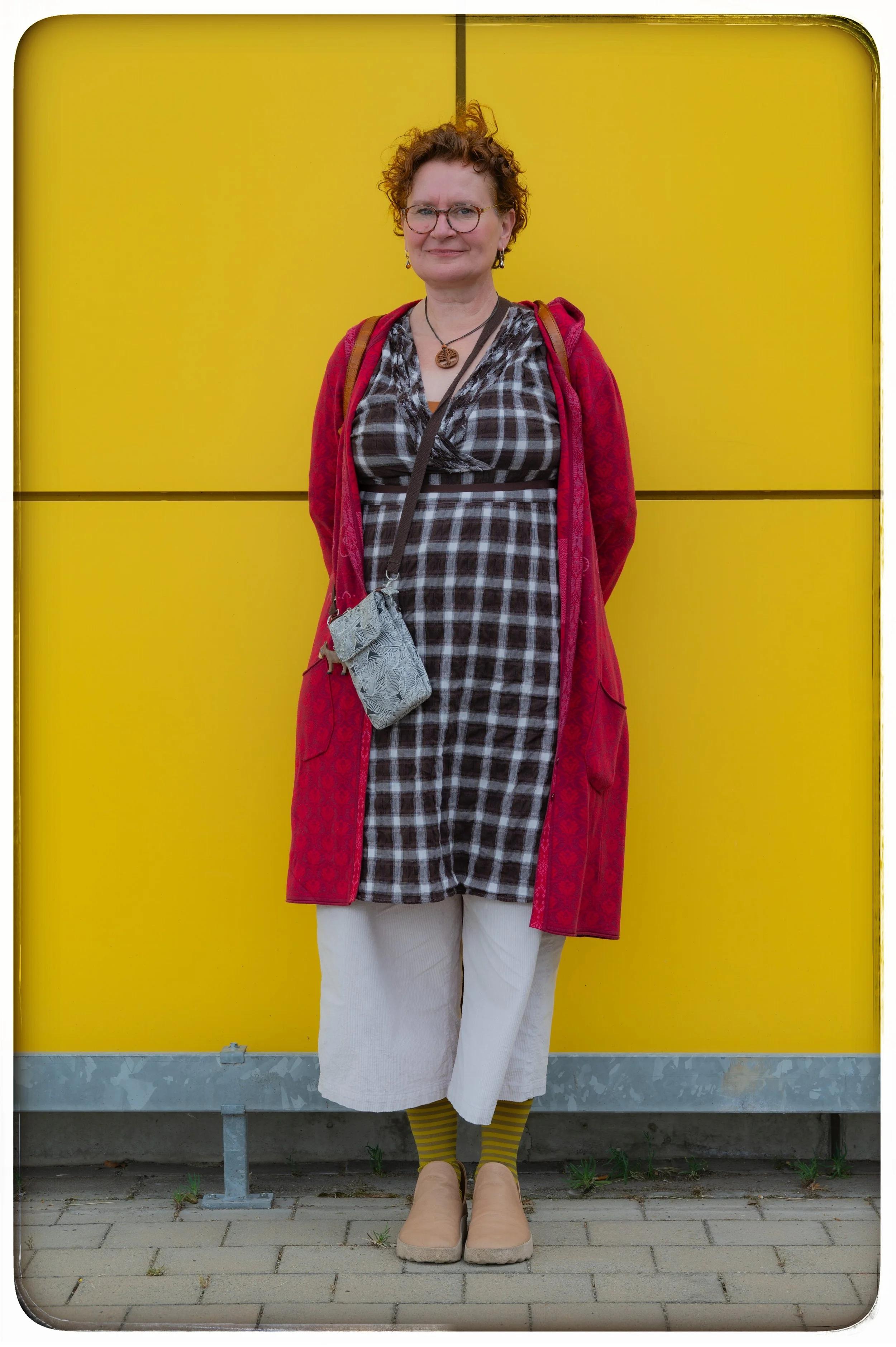 Frau steht vor einer gelben Wand, trägt ein rotes Mantel, ein schwarz-weiß kariertes Kleid, weiße Hosen, grün-gelbe Socken und beiger Schuhe. Sie hat kurze, lockige Haare, trägt Brille und ein Schmuck. Sie hat eine kleine graue Tasche um den Hals.