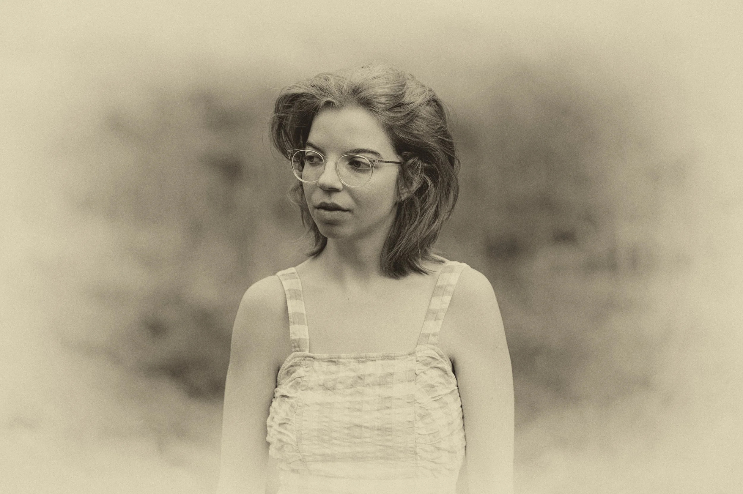 Junge Frau mit Brille und lockigem Haar, trägheit eines Sommerkleids, blickt nach links, im Hintergrund unscharfe Naturaufnahme.