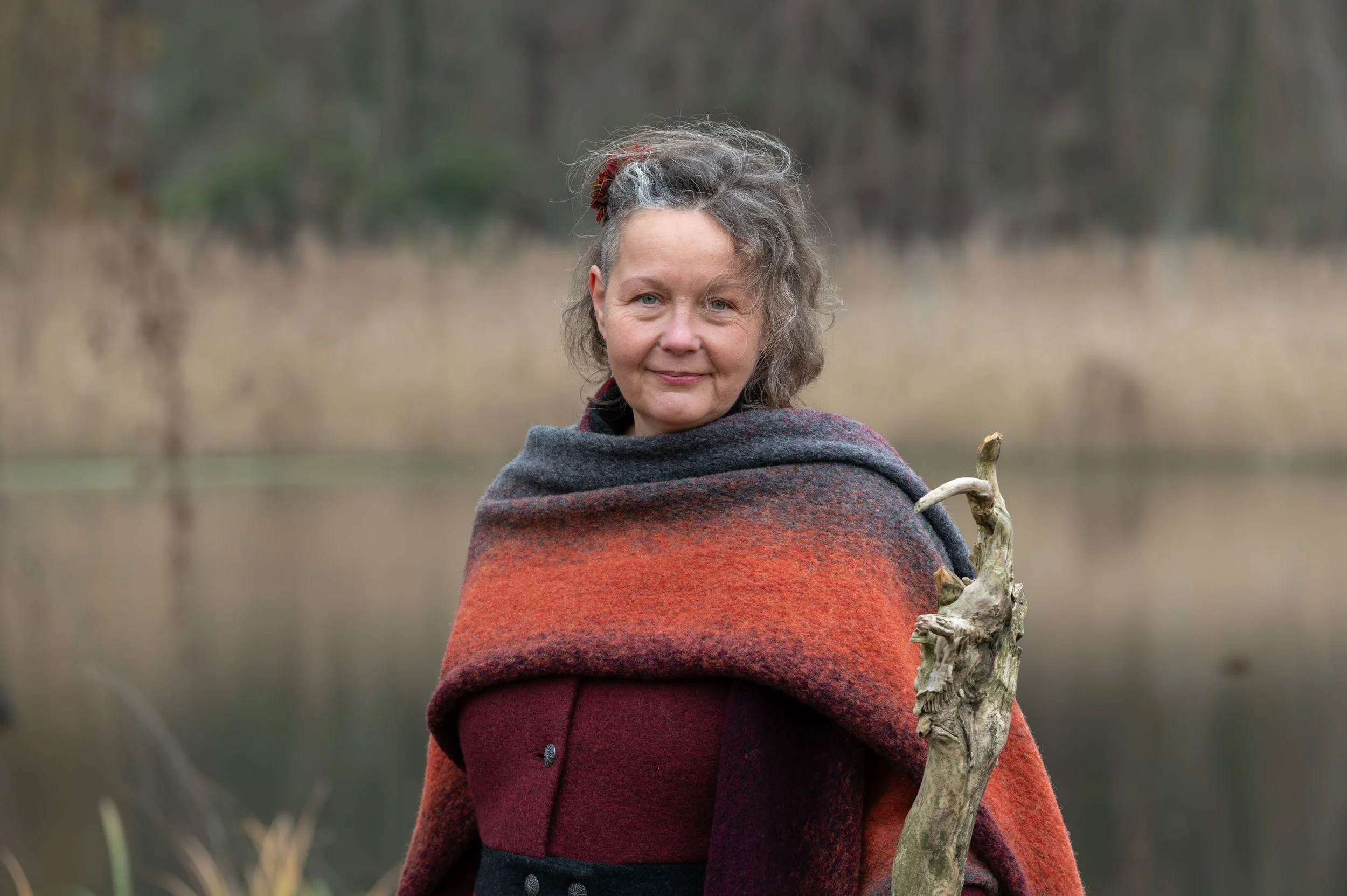 Eine ältere Frau mit grauem, gewelltem Haar trägt eine rot-schwarz-graue Schalkragenjacke und steht vor einem See in einer natürlichen Umgebung, hält einen knorrigen Ast in der Hand.