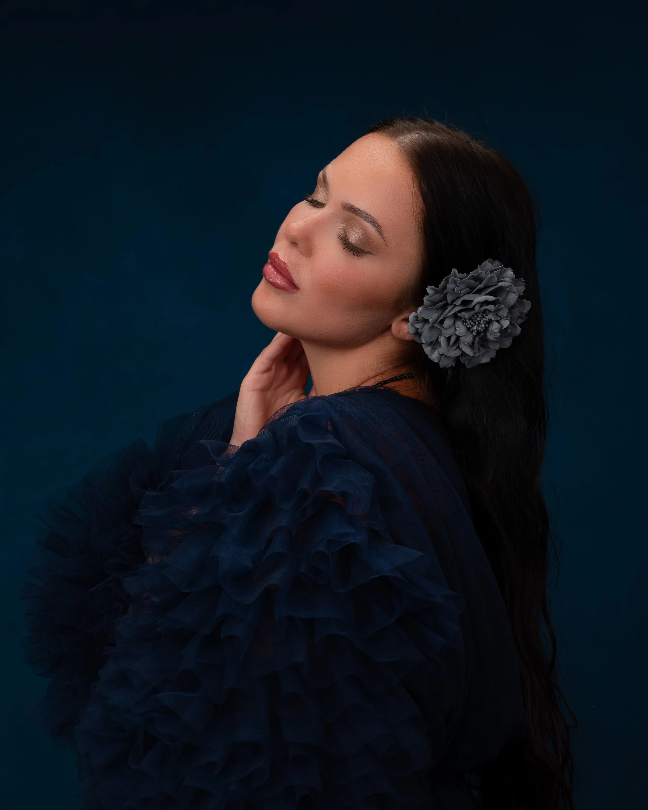 Porträt einer Frau mit dunklem Haar und einem großen grauen Hortensienblume im Haar, mit geschlossenen Augen vor dunkelblauem Hintergrund.