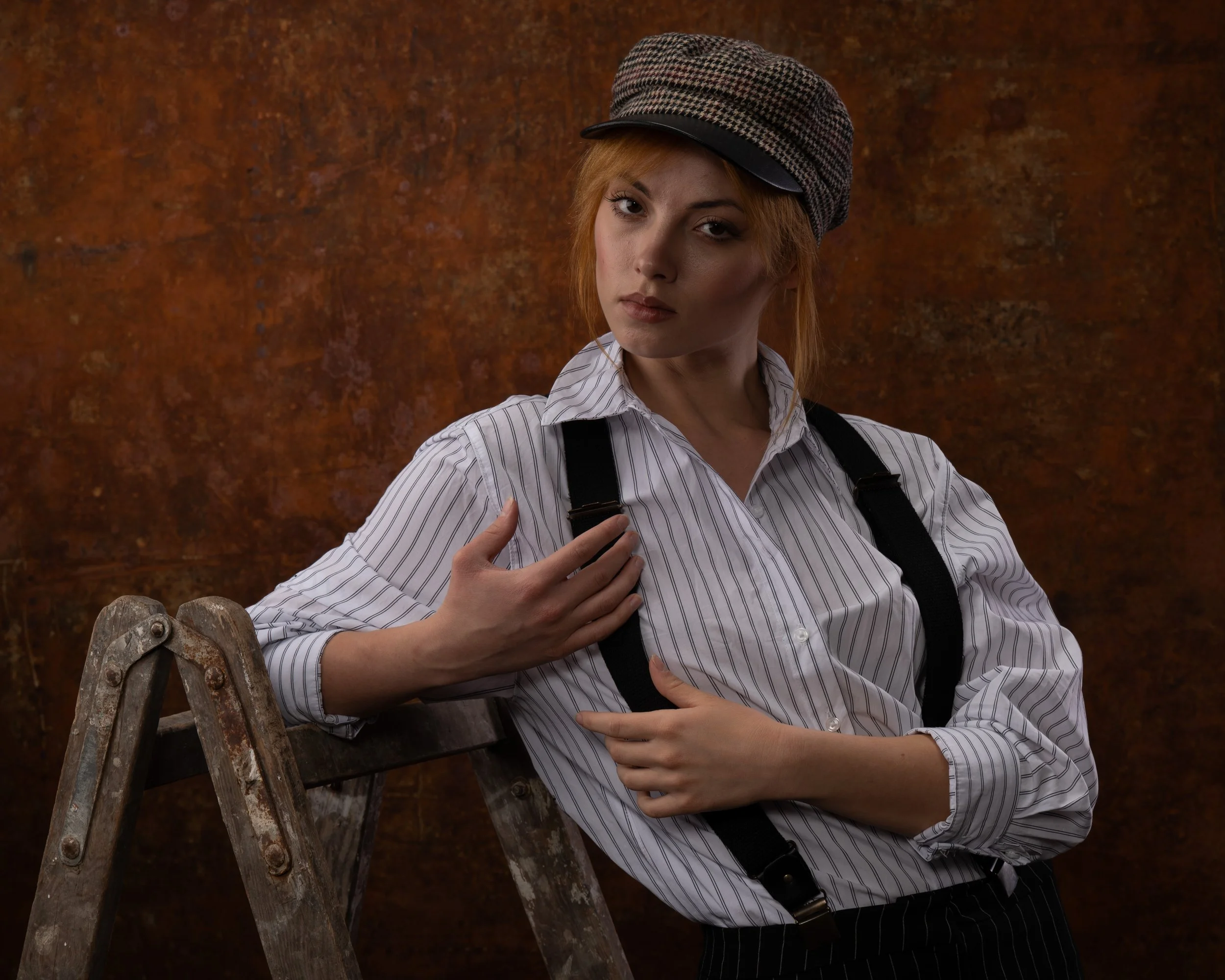 Junge Frau mit rotem Haar, Kappe und gestreiftem Hemd, steht neben einer Leiter, vor einem rustikalen, braunen Hintergrund.
