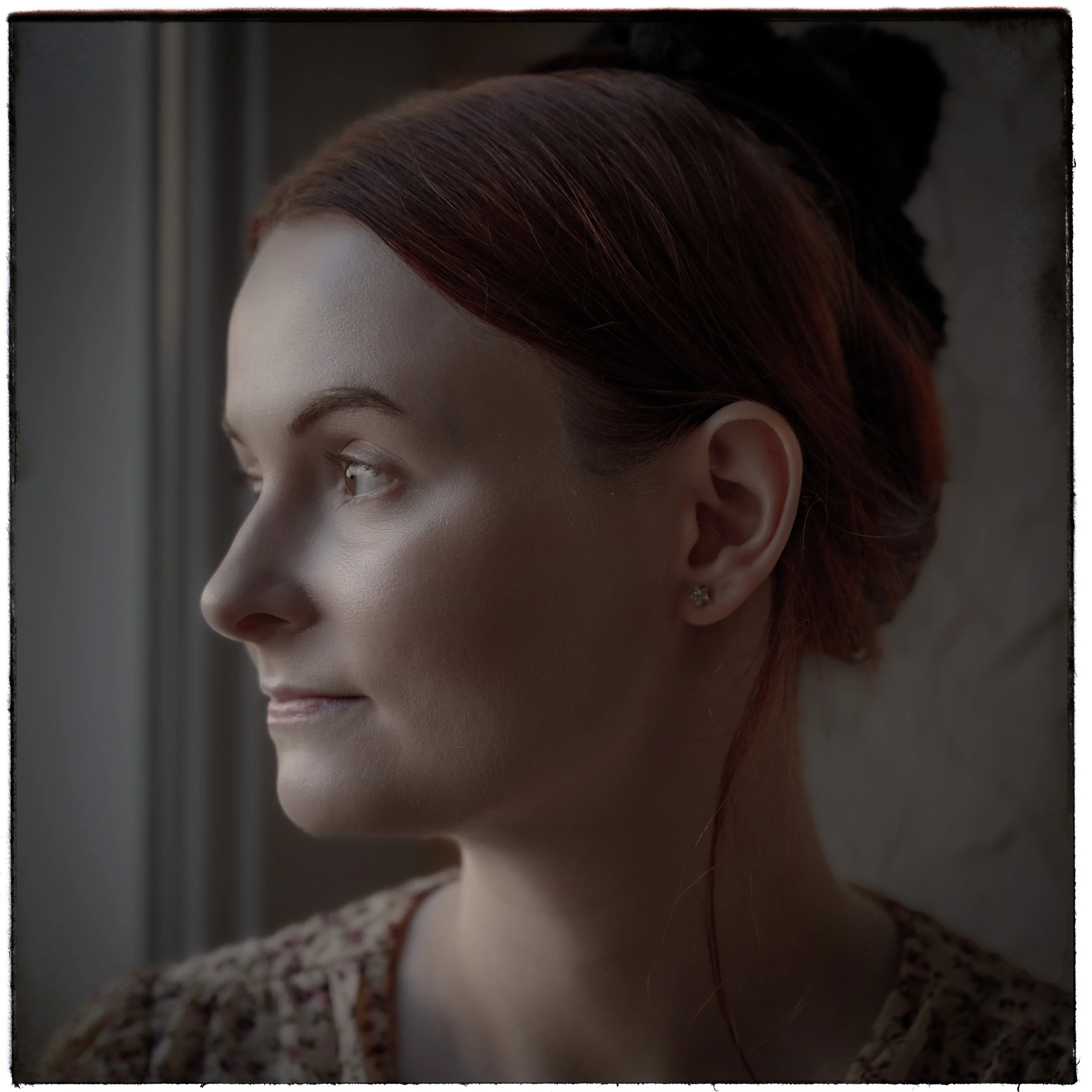 Porträt einer jungen Frau mit rotem Haar, die am Fenster steht und nach draußen schaut.