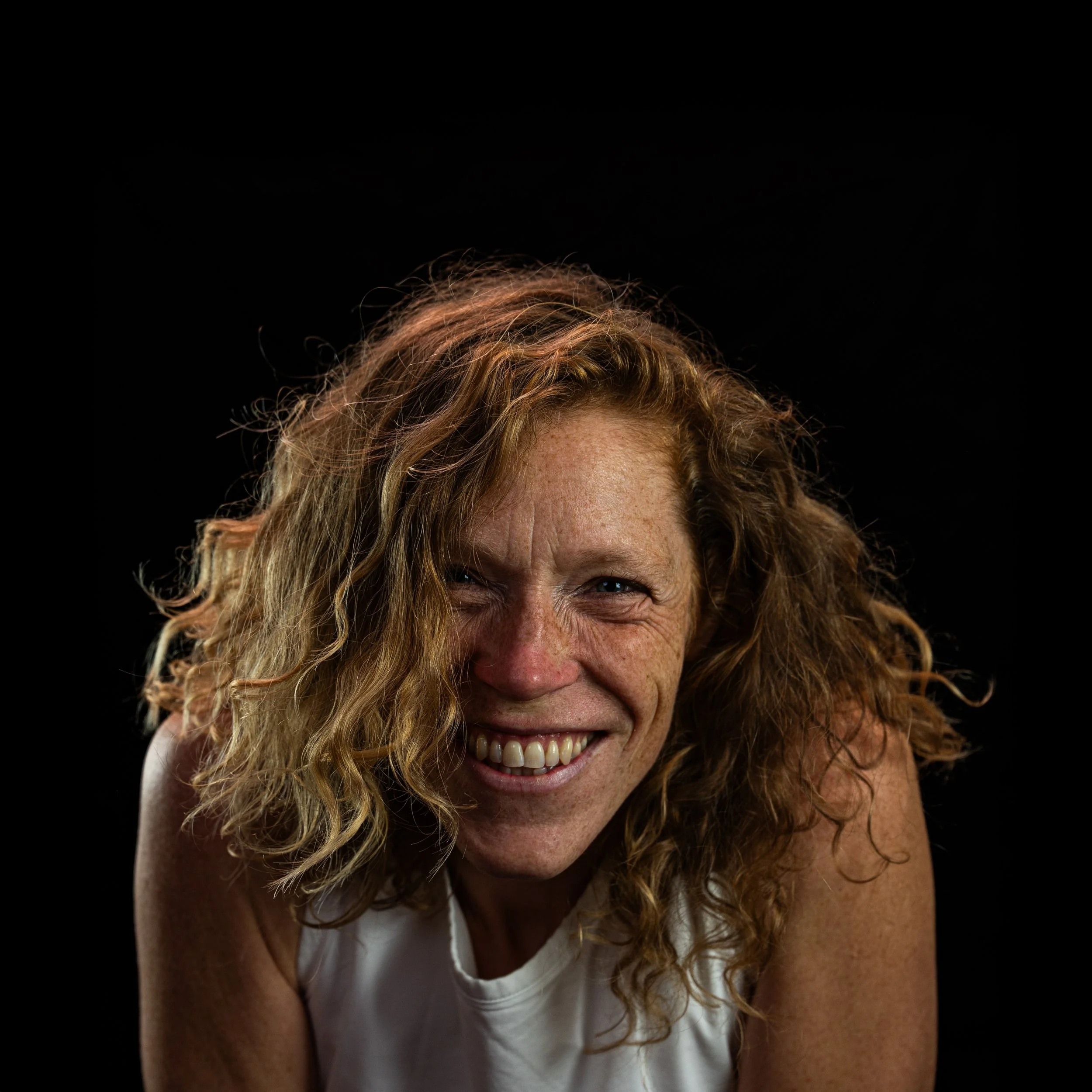 Ein Porträt einer lächelnden Frau mit roten, lockigen Haaren und Sommersprossen vor einem schwarzen Hintergrund.