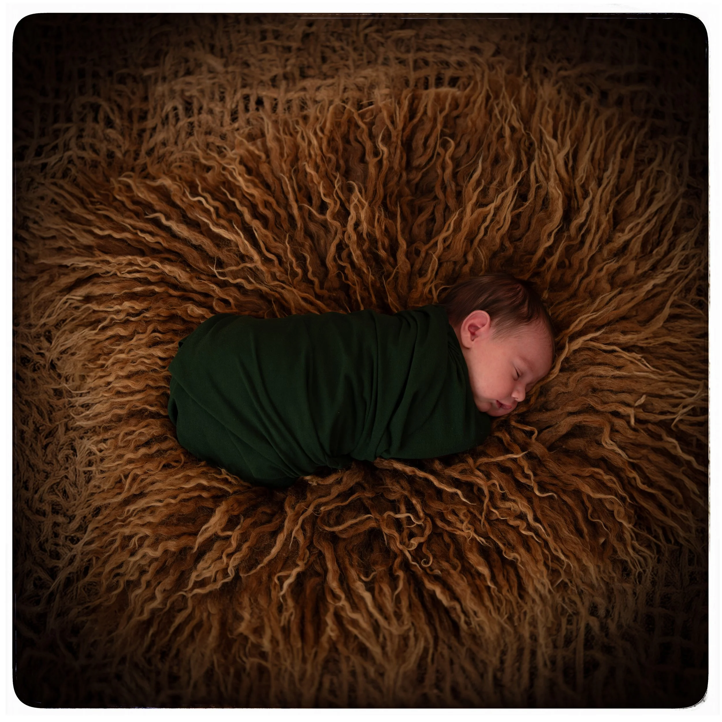 Ein schlafender Säugling, der in einer grünen Kleidung eingewickelt ist, liegt auf einem großen, braunen, flauschigen Teppich.