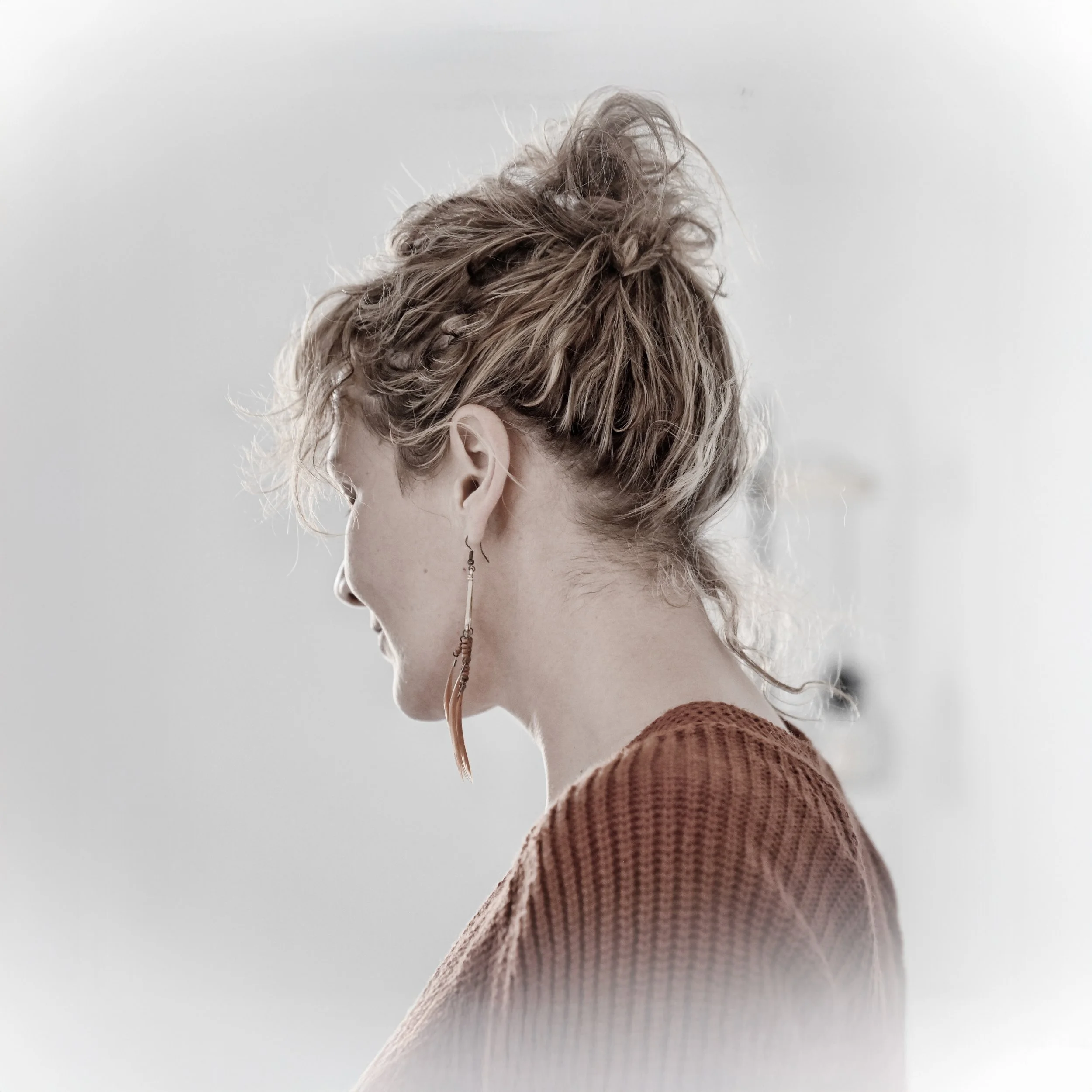 Porträt einer lachenden Frau mit wirrem, braunem Haar, die eine lange Ohrring trägt, im Profil aufgenommen, trägt ein braunes gestricktes Oberteil, Hintergrund ist unscharf und hell.