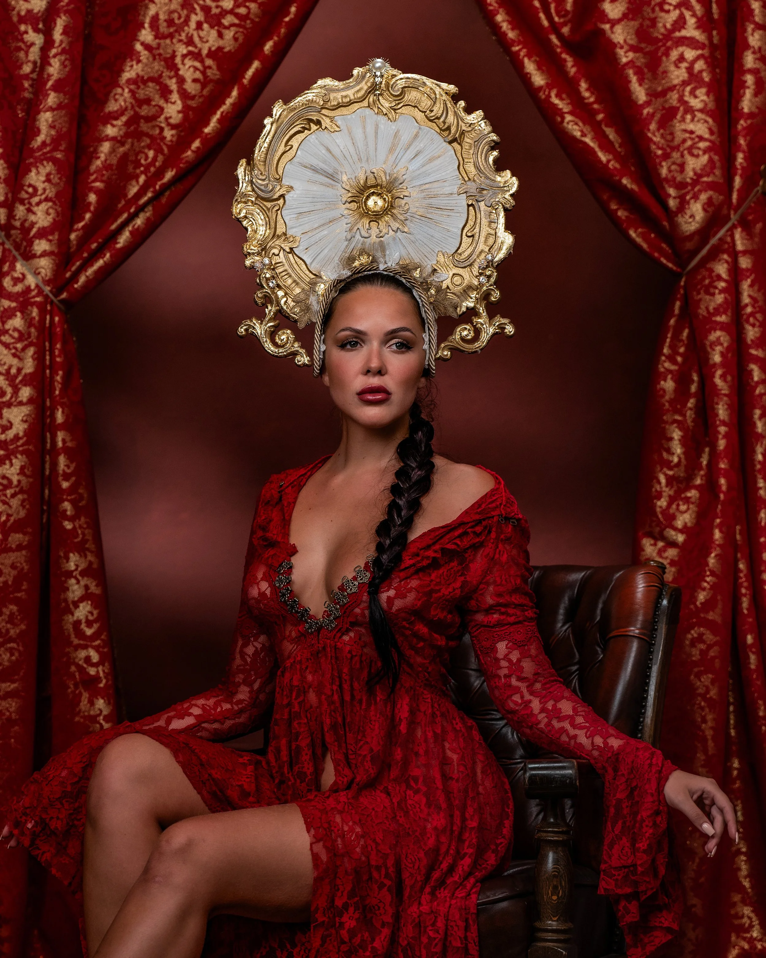 Frau in rotem Spitzenkleid sitzt auf einem Sessel vor roten Samtvorhängen, trägt ein großes, goldverziertes Heiligenschein-ähnliches Kopfschmuck und hat lange dunkle Haare in Zopf.
