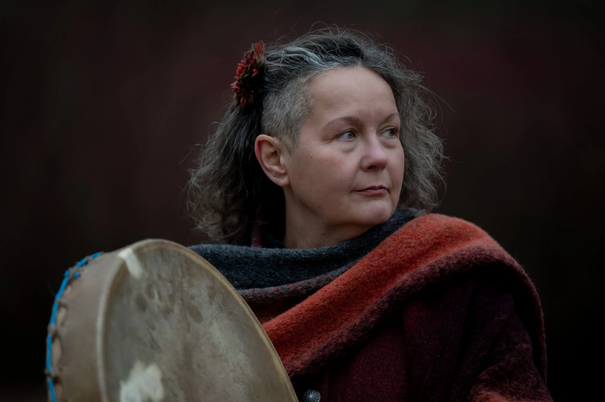 Eine Frau mit grauen, lockigen Haaren, die teilweise kurz an den Seiten sind, trägt einen dunkelroten und schwarzen Schal und hält ein rundes, handgefertigtes Trommelinstrument. Der Hintergrund ist dunkel und unscharf.