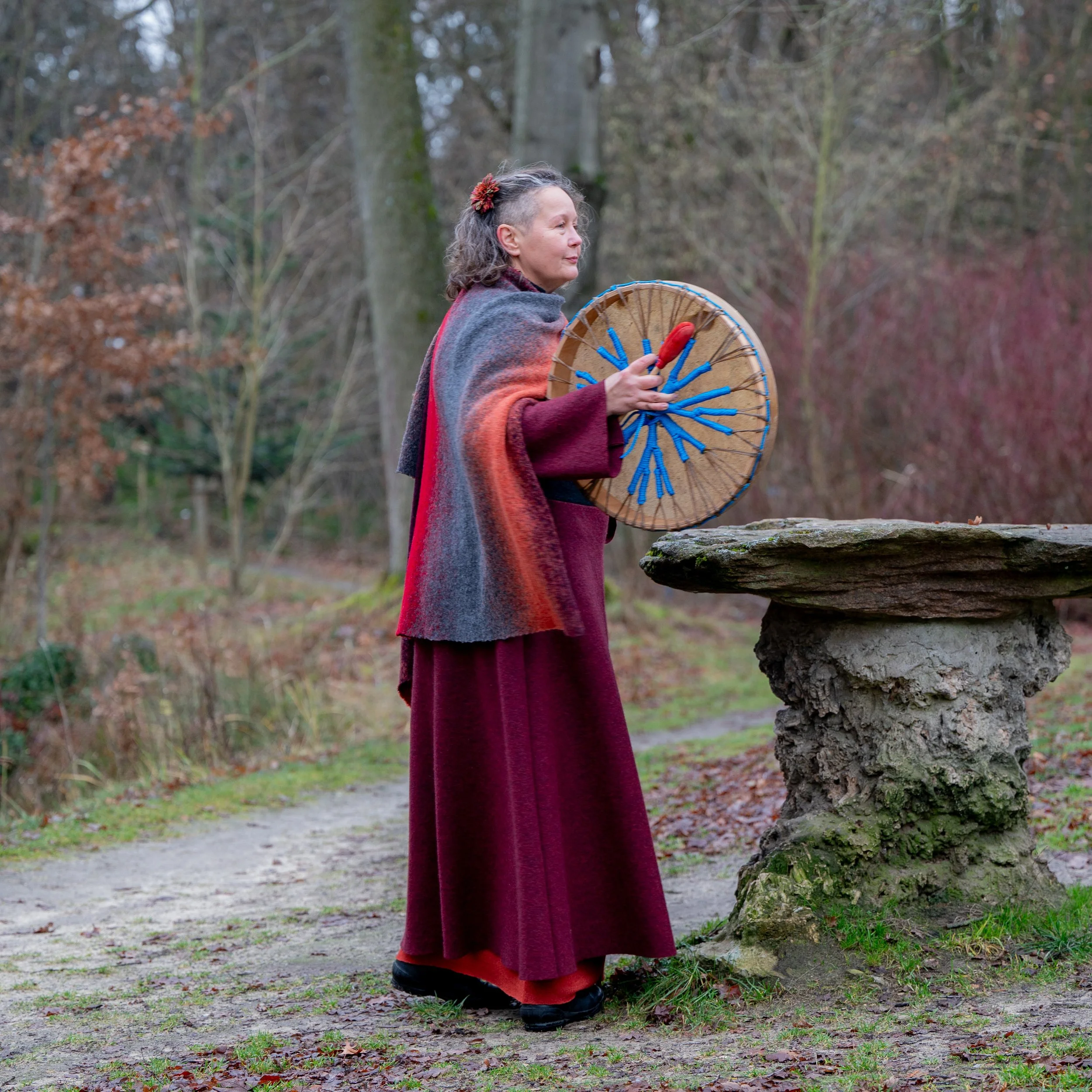 Eine ältere Frau in einem langen roten Kleid und einem grauen, farbenfrohen Schal steht im Wald, hält einen runden, dekorativen Regenschirm mit blauen Schnüren und einer roten Spitze, während sie neben einem alten Stein-Tisch steht.