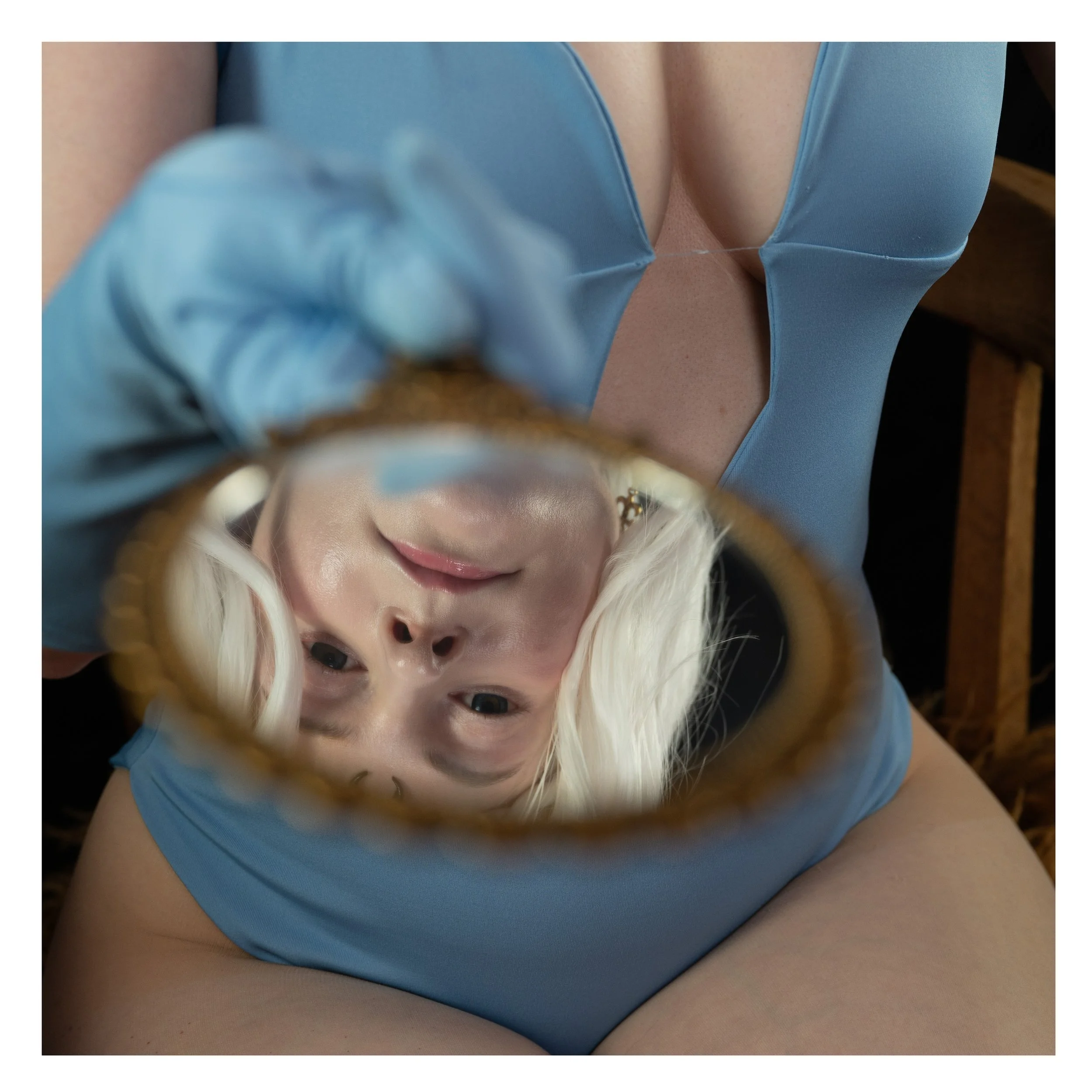 Frau mit blonder Haar, die in einem blauen Kleid im Spiegel schaut, von unten fotografiert, während sie das Glas hält.